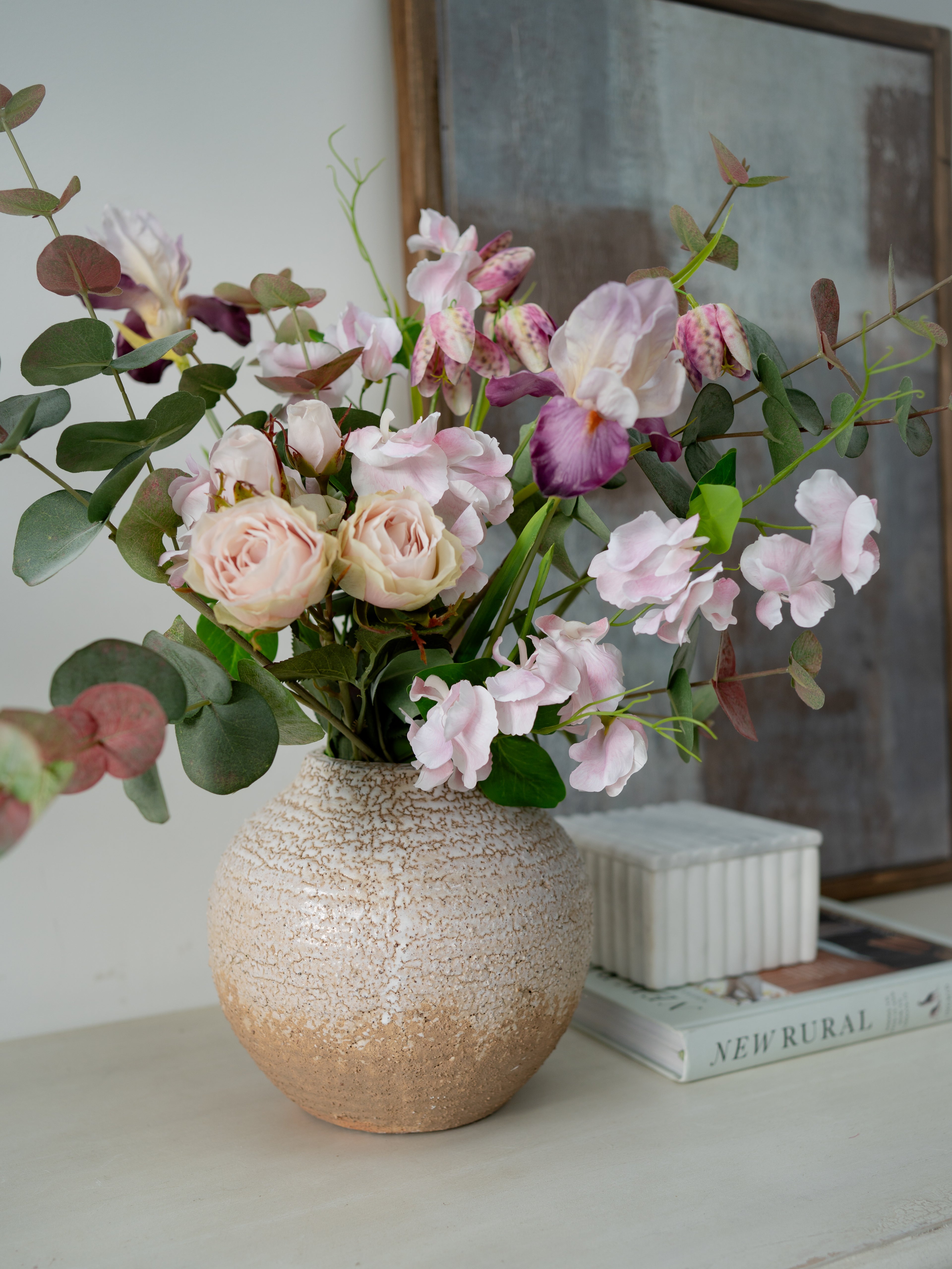 Vase with artificial pink flowers on a surface with books and a framed picture in the background