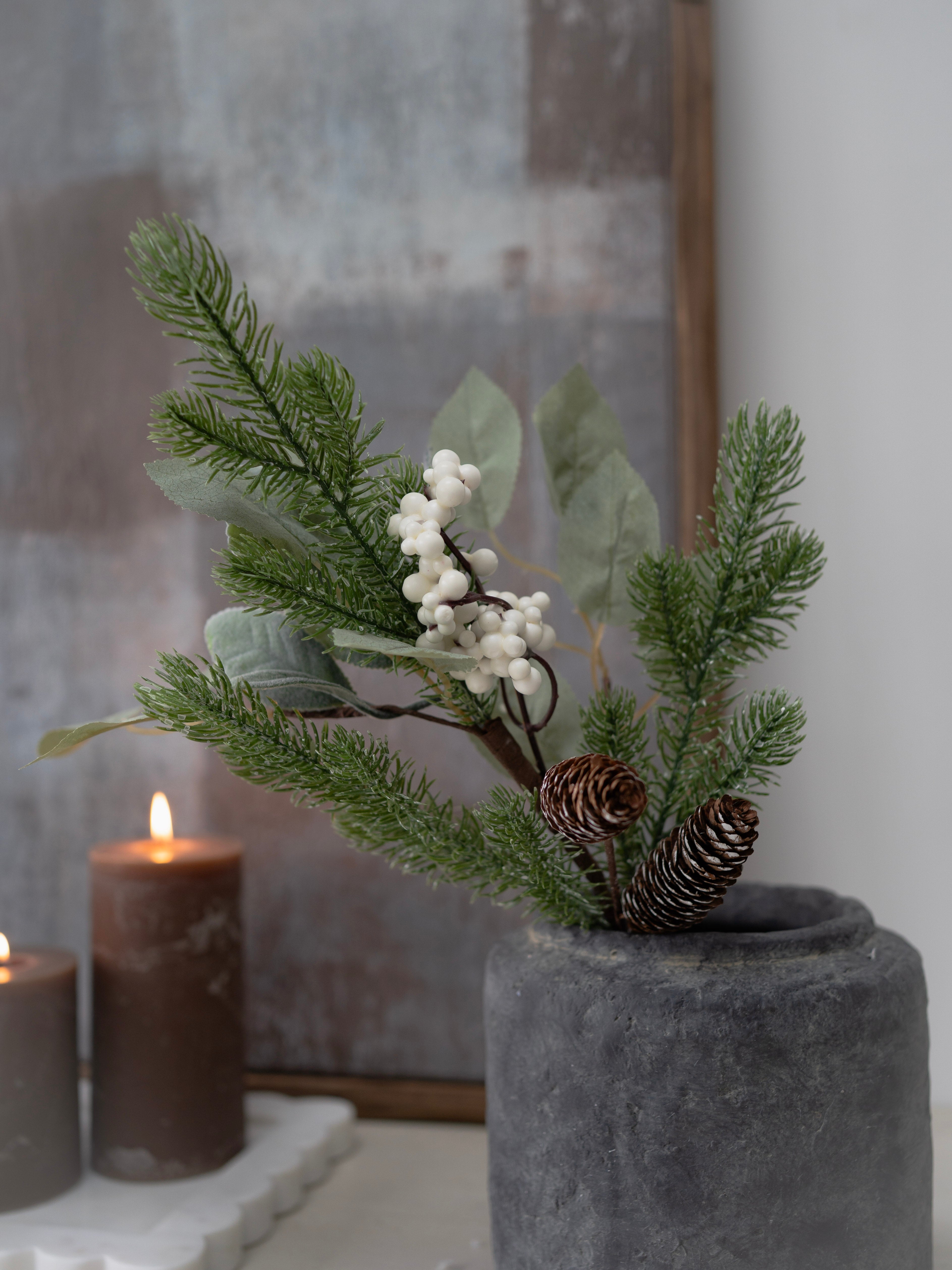 Decorative plant arrangement with berries and pinecones in a pot, next to a candle on a marble surface.