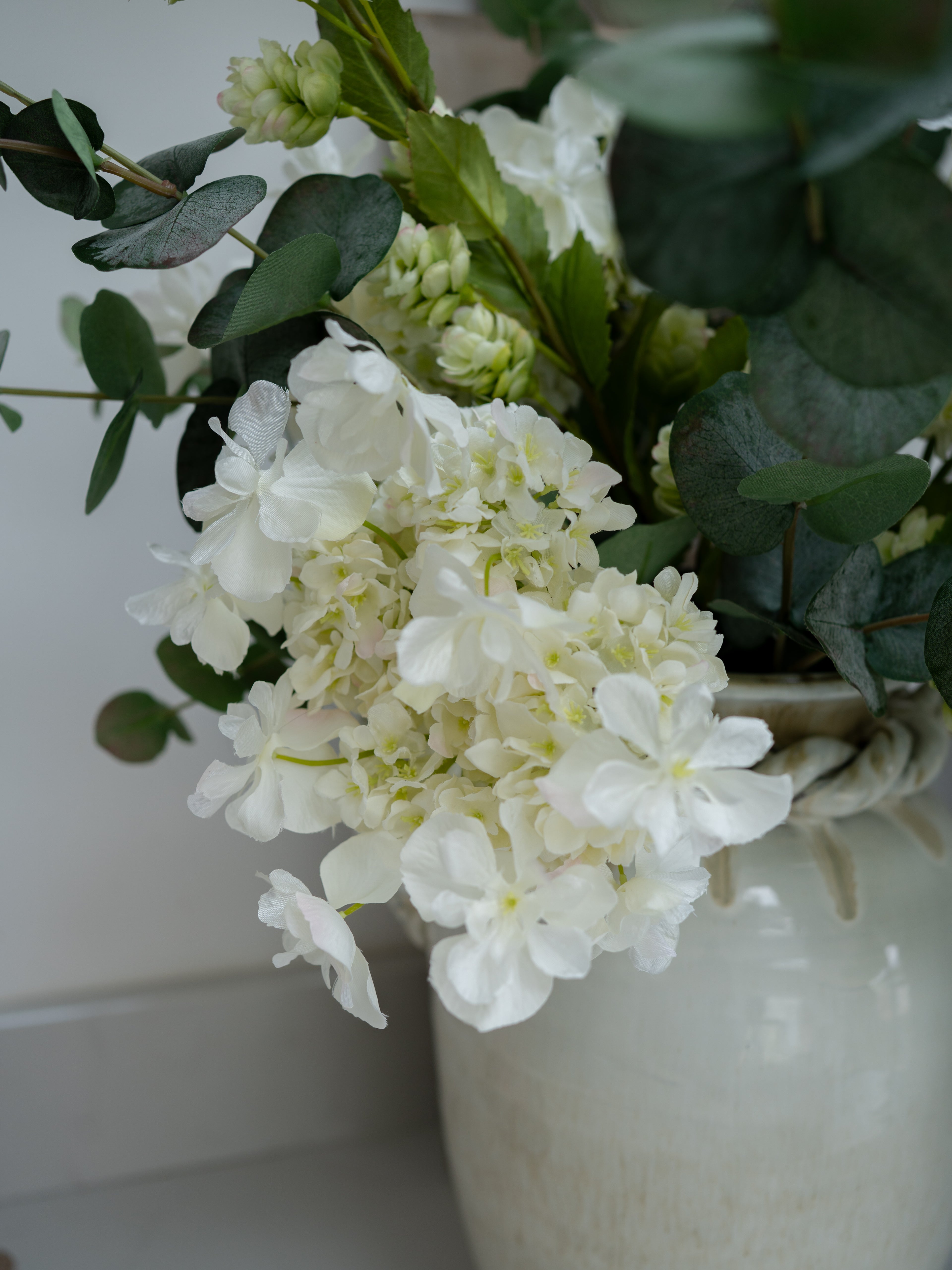 White faux hydrangeas flowers in a vase with green leaves on a white background