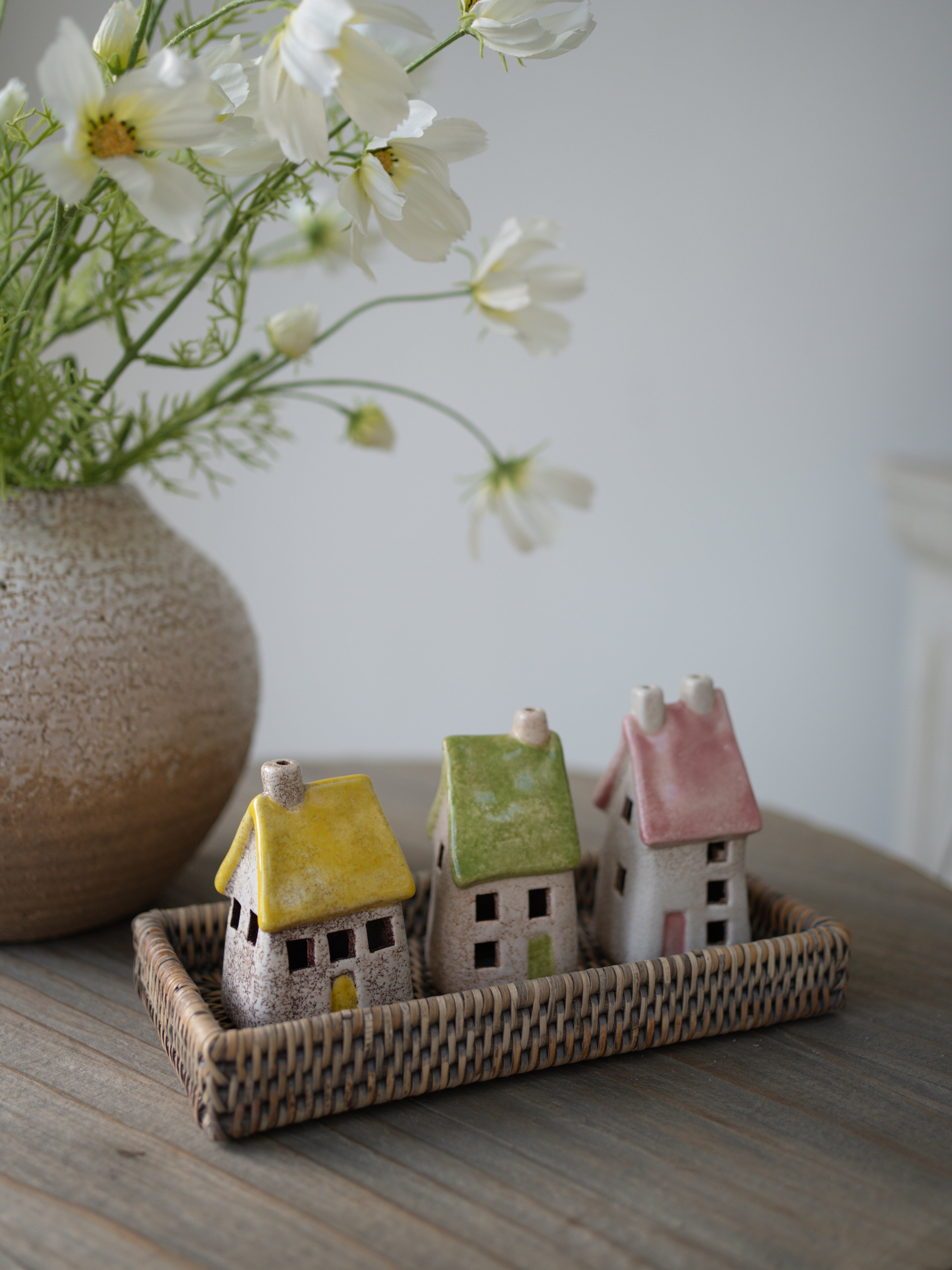 pastel toned roof stoneware houses on grey washed rattan tray with white faux cosmos and vase in the background on grey wooden table