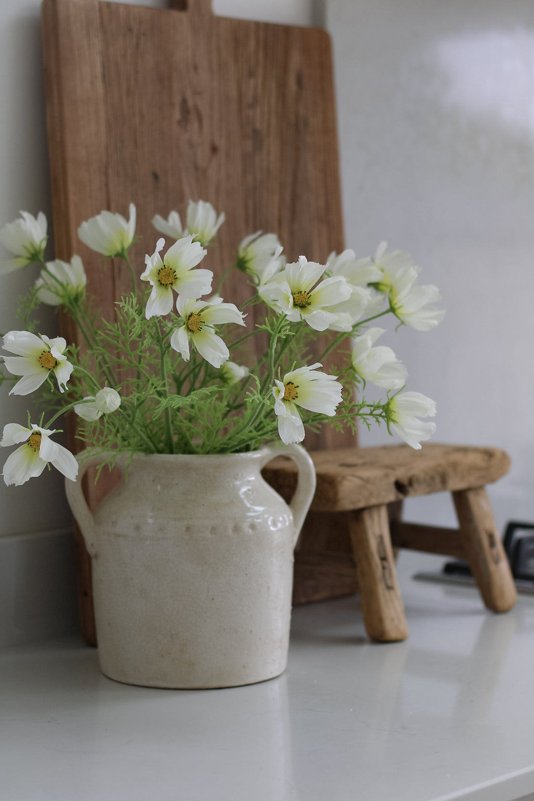 faux White cosmos flowers in a ceramic vase on a white surface with a wooden cutting board in the background.
