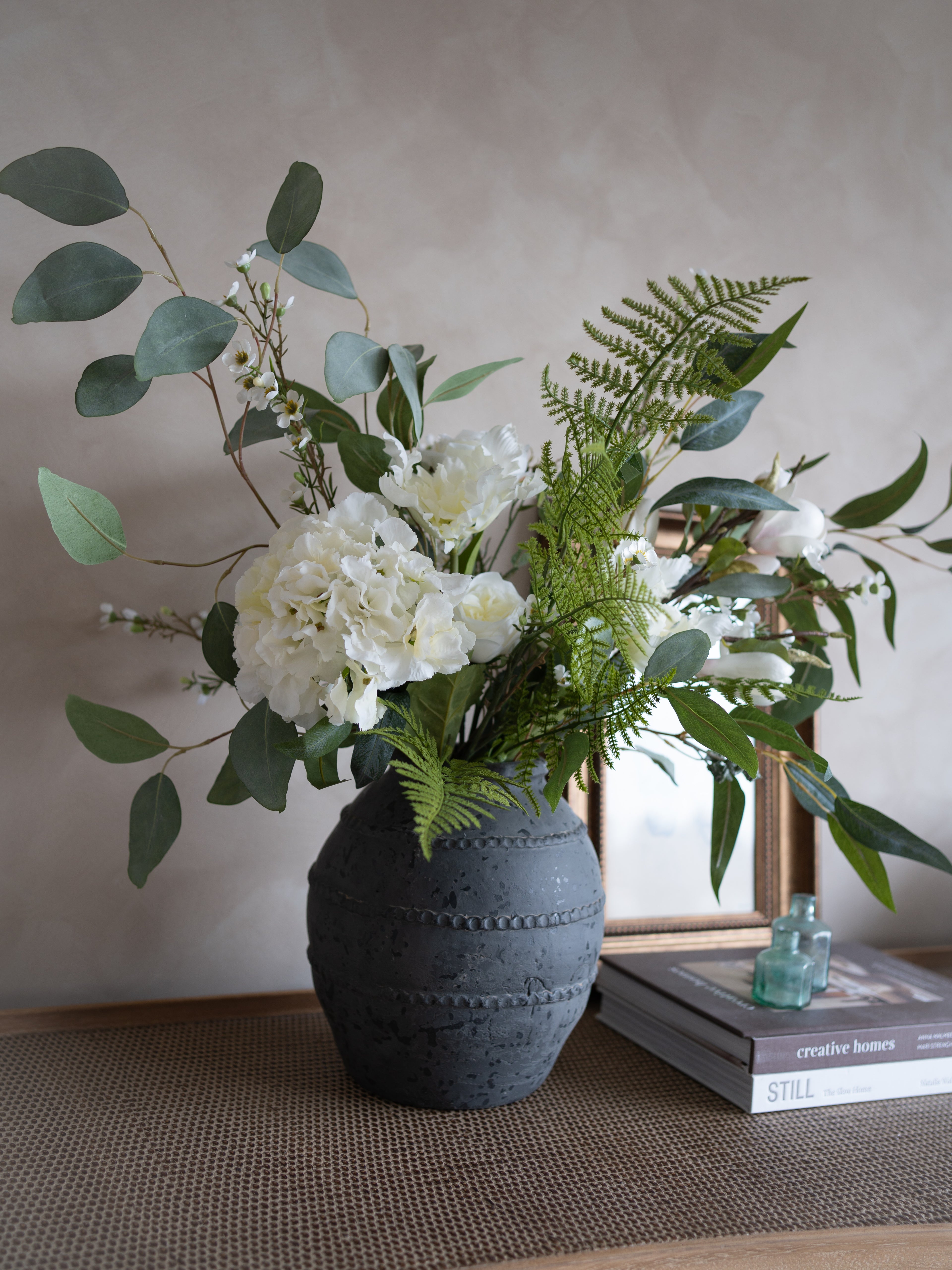 Decorative arrangement of artificial white flowers and greenery in a textured black vase on a table.