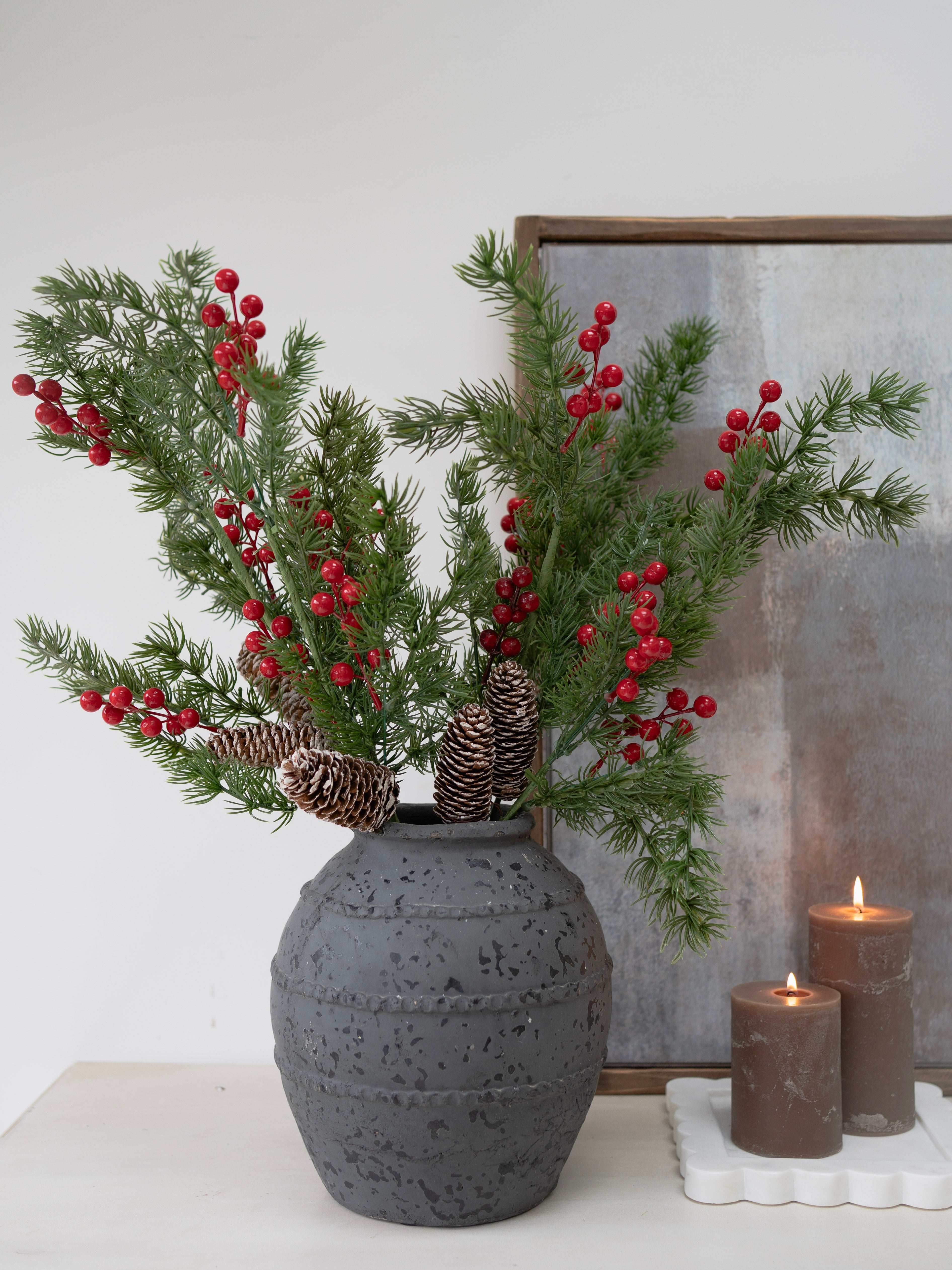 Decorative arrangement with faux christmas green branches and red berries in a textured gray vase on a white surface.