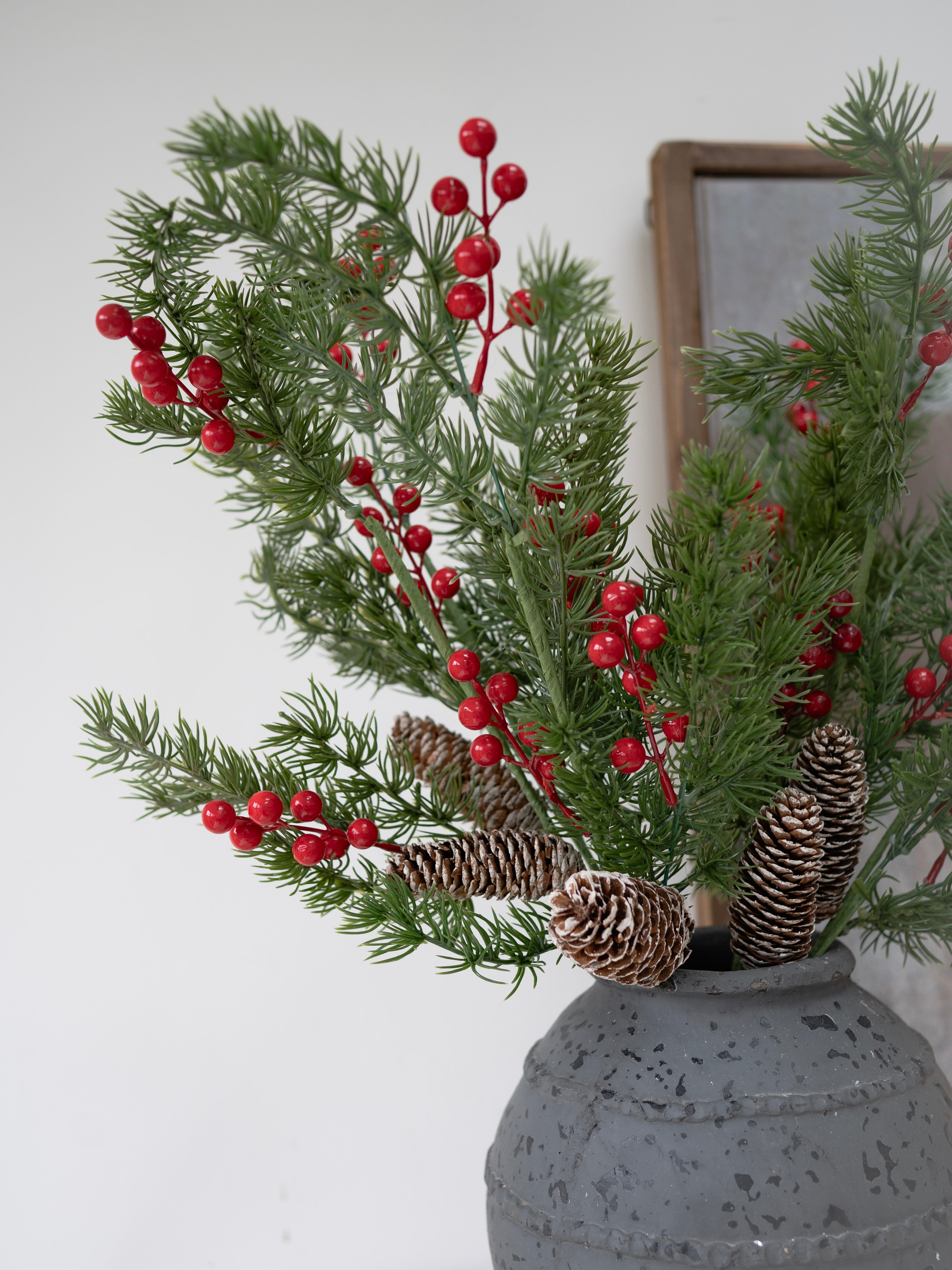 Decorative arrangement with faux christmas greenery, red berries, and pinecones in a gray vase against a white wall.