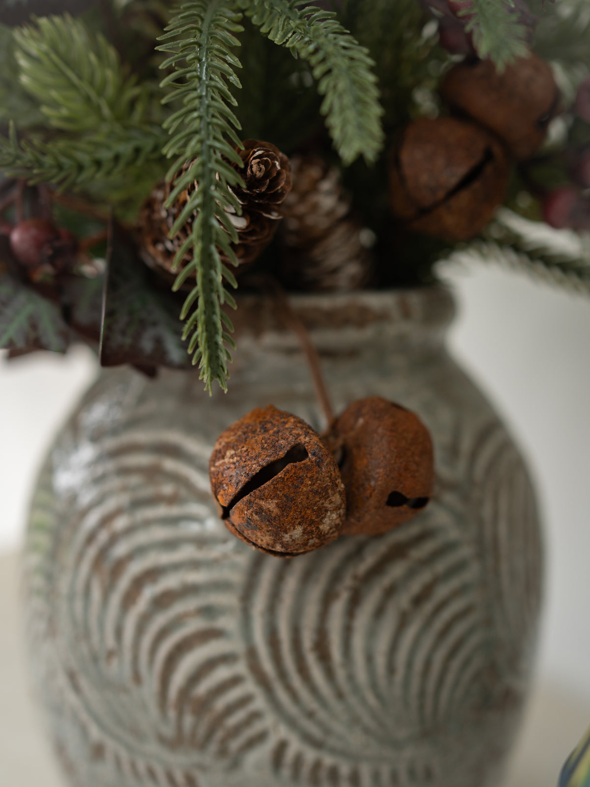 Rustic christmas floral arrangement featuring two brown jingle bells, pinecones, and green pine needles in a textured gray ceramic vase.