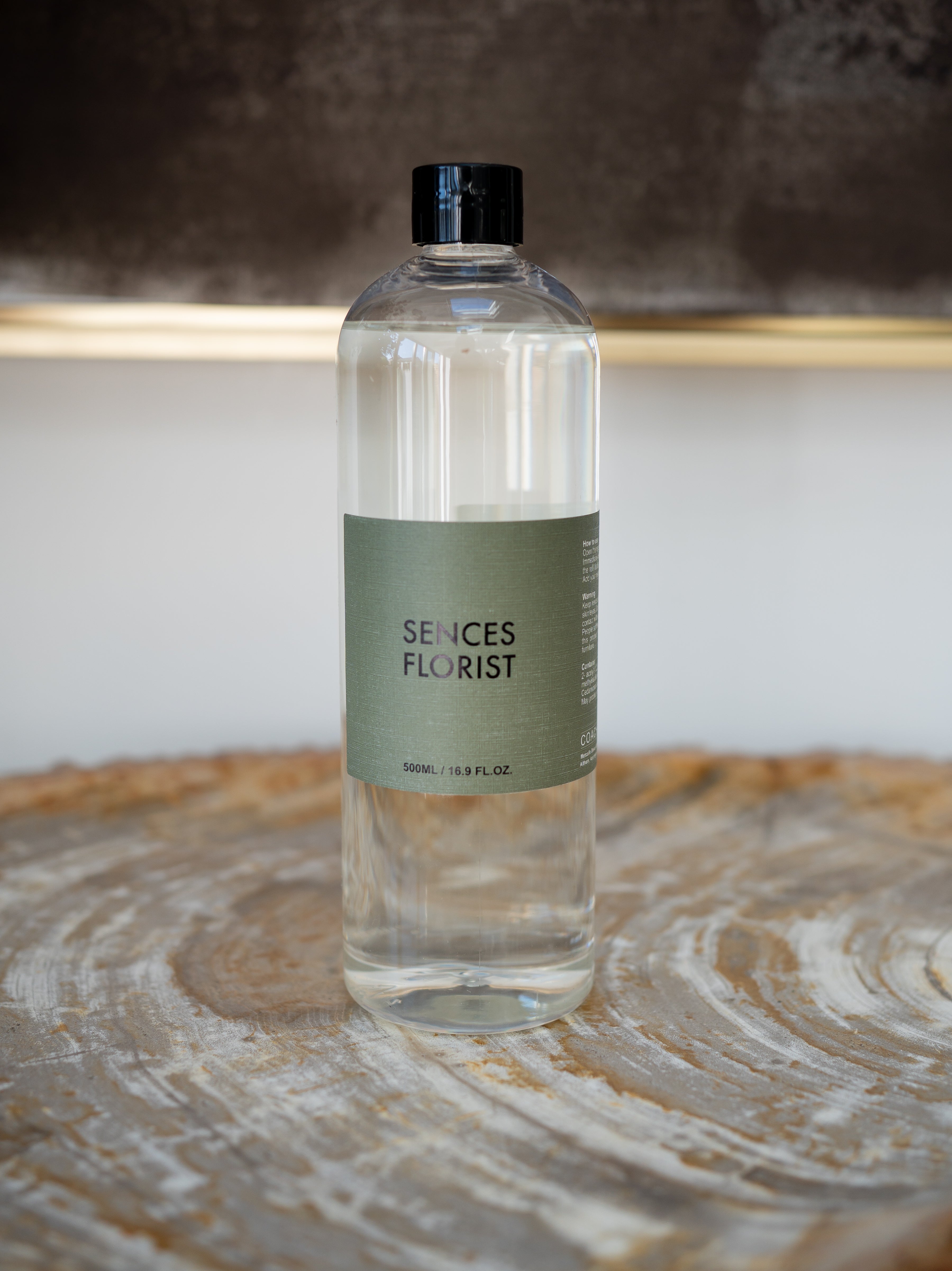 A clear glass bottle of Scentes Florist refill bottle with a black cap and a muted sage green label, resting on a rustic wood slice surface.