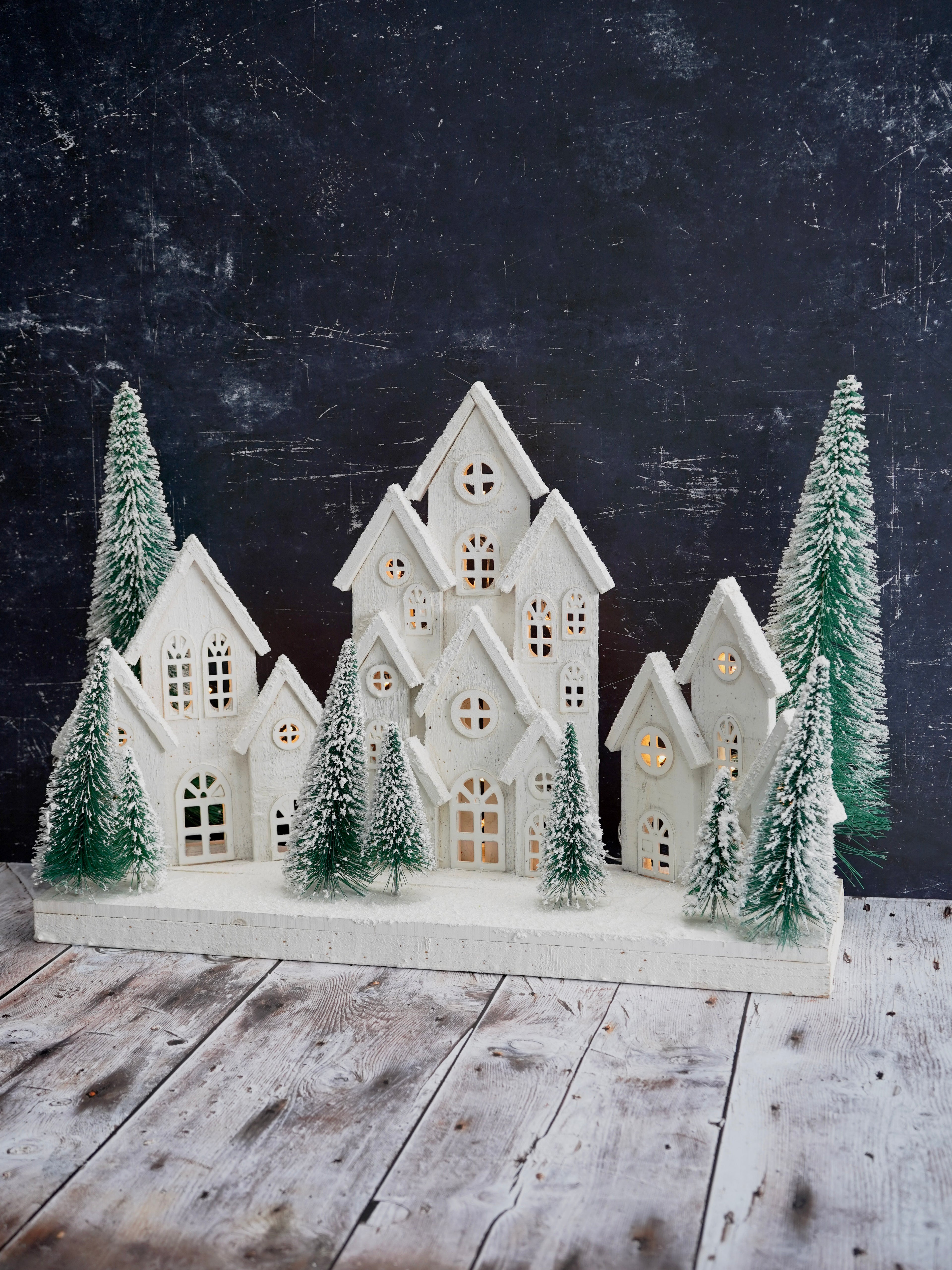 Decorative model of a snowy village with houses and trees on a wooden surface against a dark background.