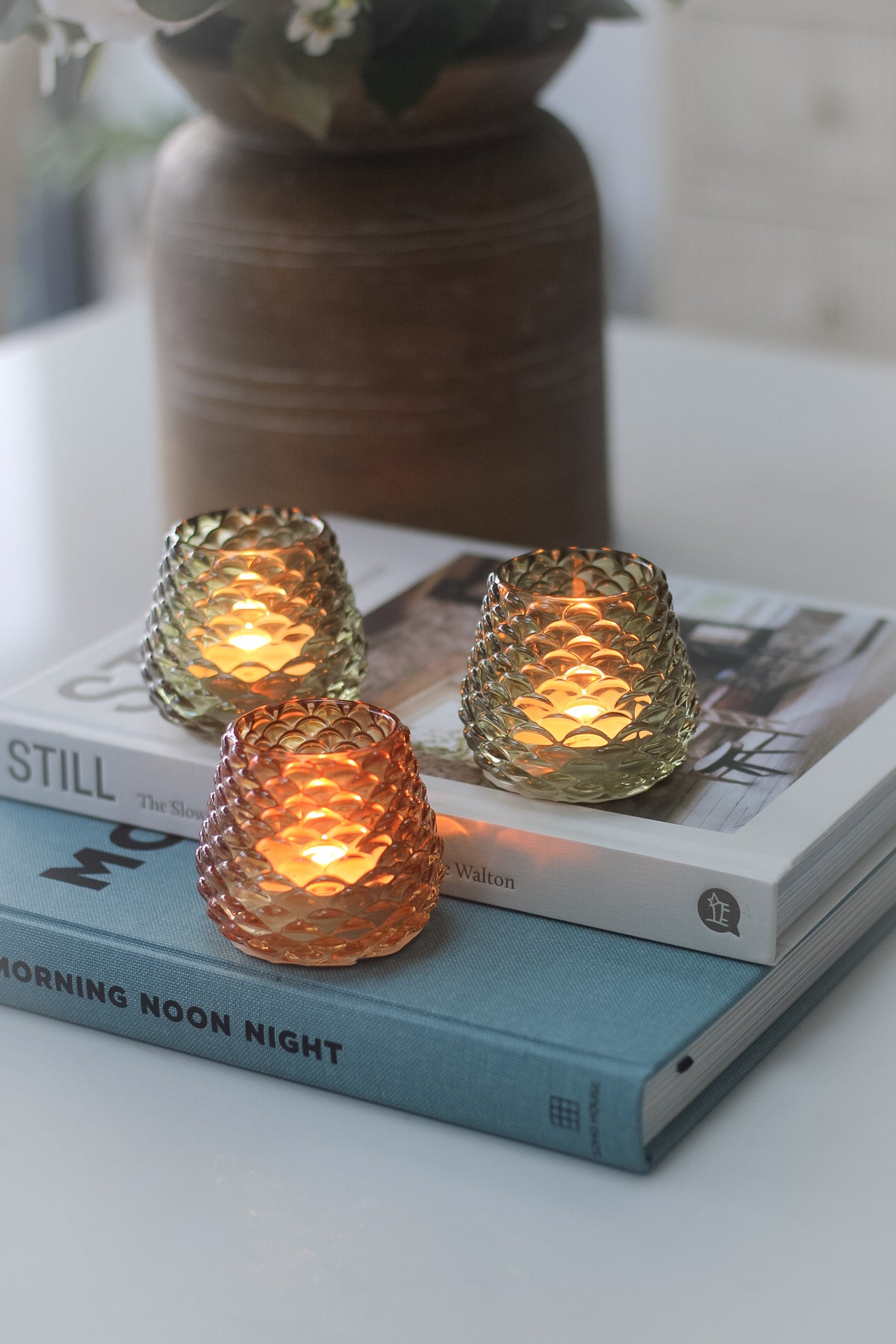 Three glass pinecone votives on a table, with one amber votive lit, placed on top of an open book.