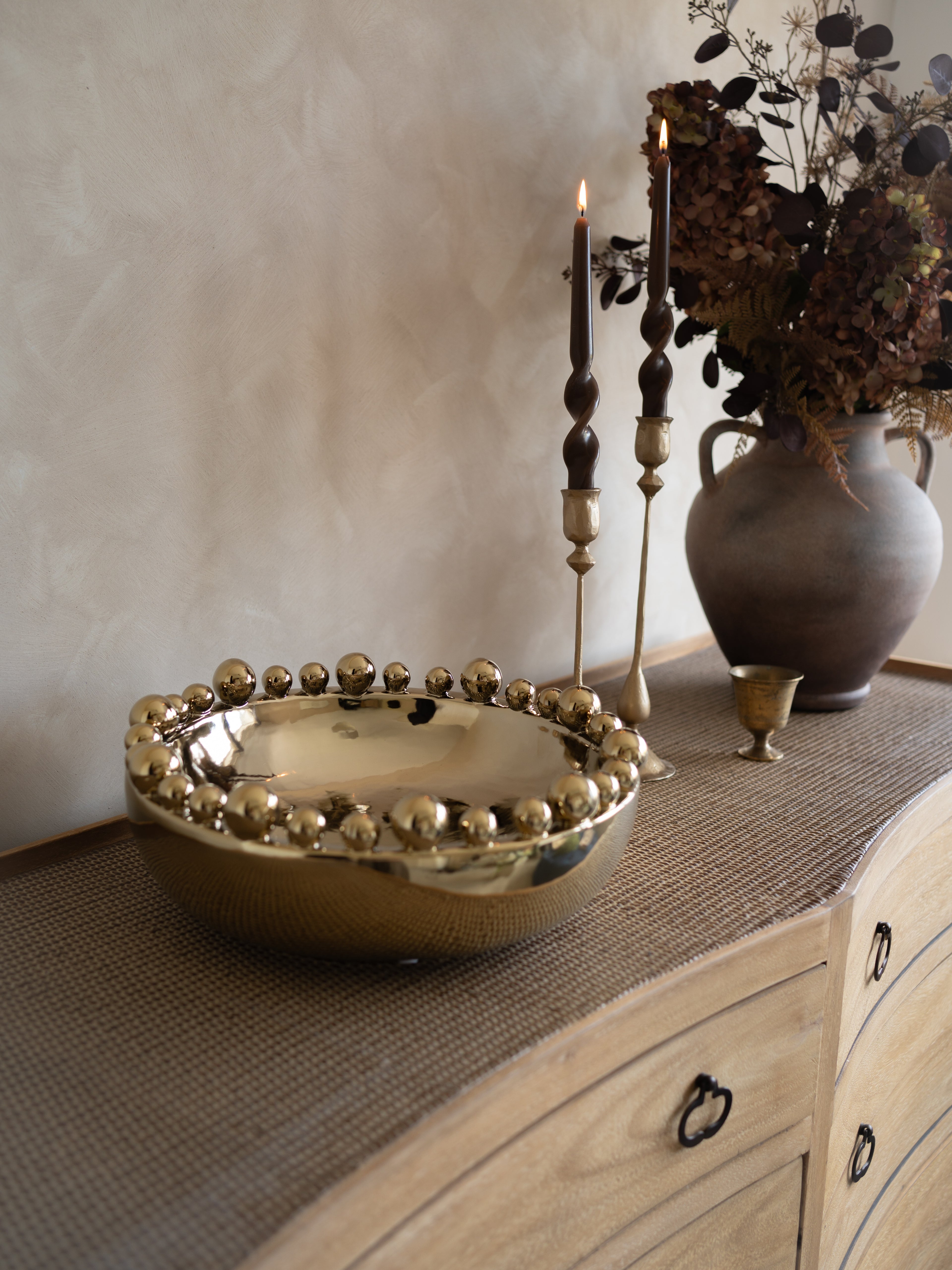 Decorative gold bowl on a wooden surface with candles and a vase in the background.