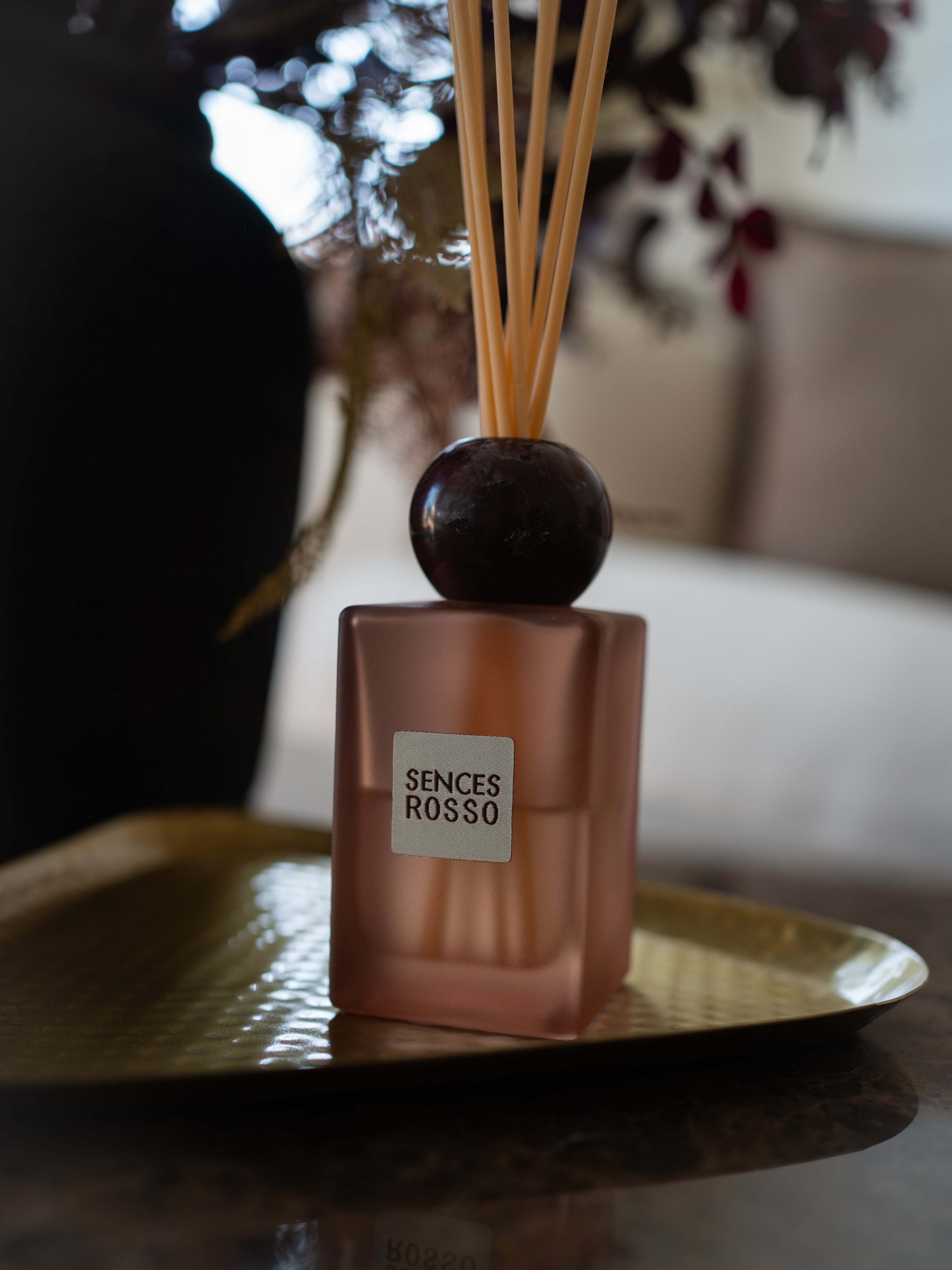 A Sences Rosso reed diffuser with a frosted rose-gold bottle, dark round stopper, and natural reeds, resting on a gold tray in a softly blurred indoor setting.