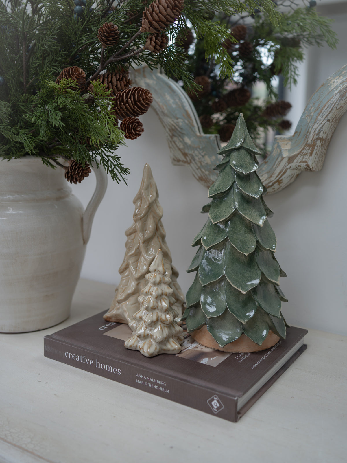 Three decorative ceramic Christmas tree ornaments, two in beige and one in green, displayed on a book next to a ceramic pitcher with pine branches.