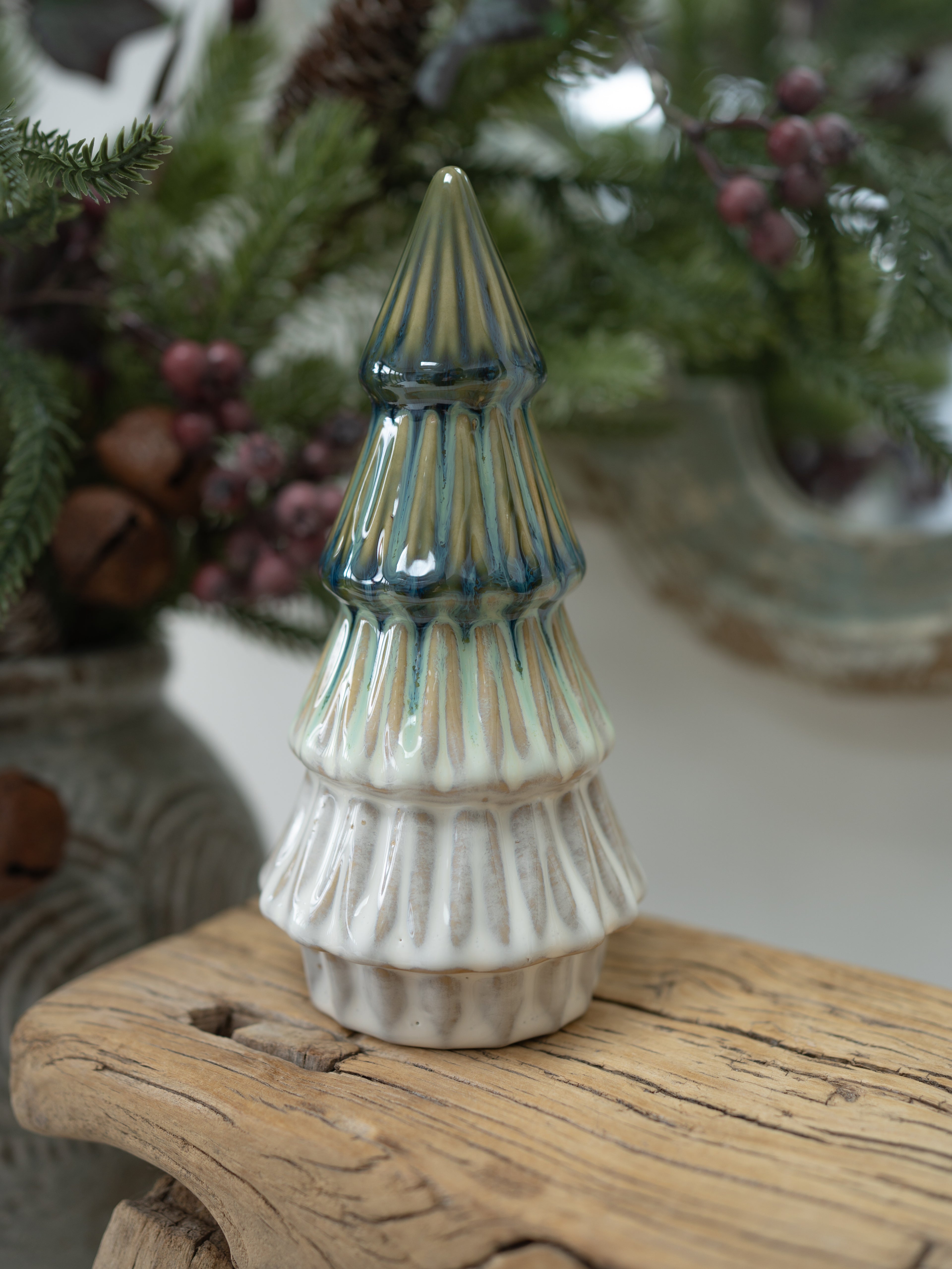 A small ceramic Christmas tree ornament with a ribbed texture, featuring a white base and a blue-green glazed top, displayed on a rustic wooden surface with a blurred holiday background.