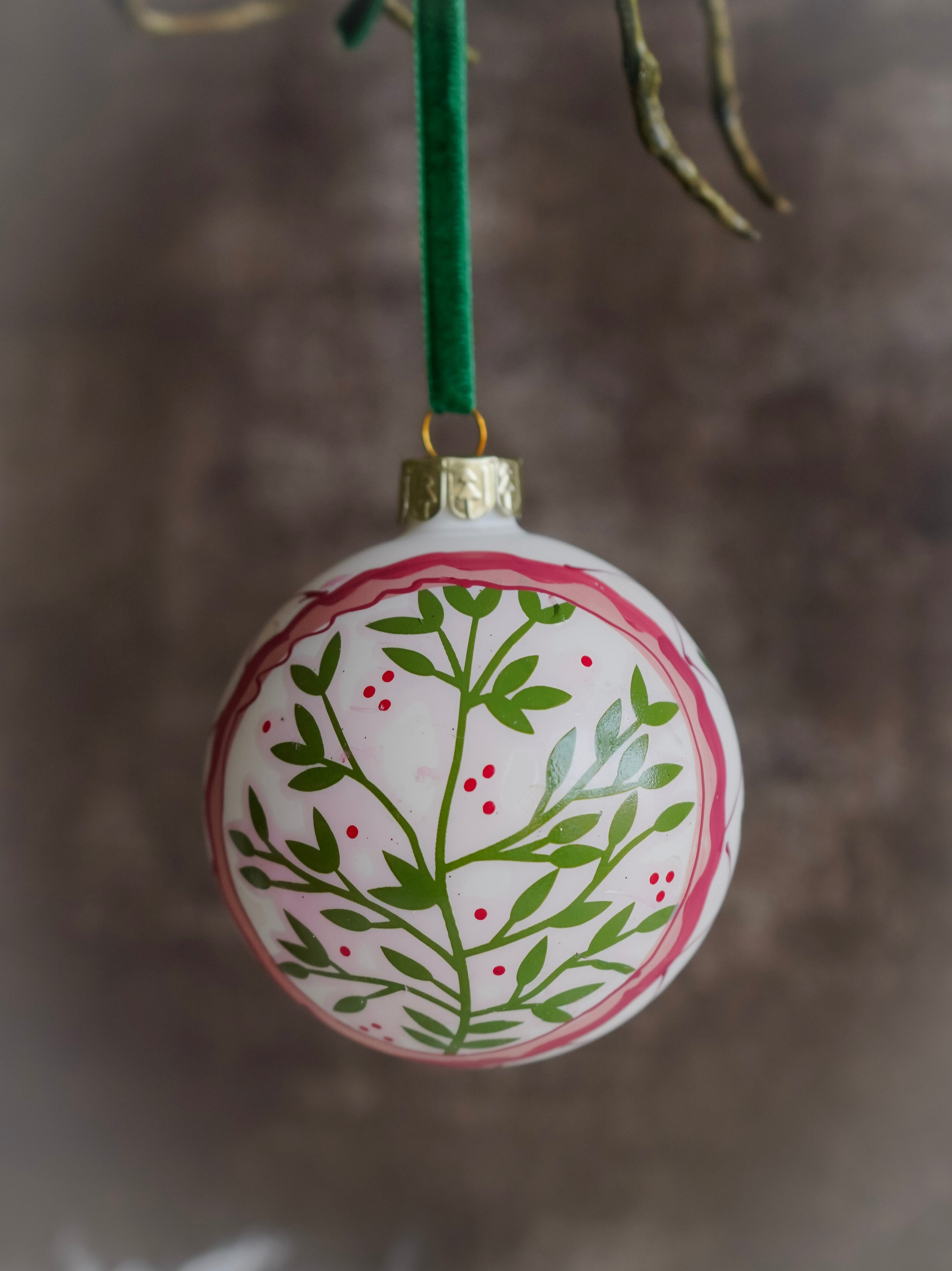 Decorative Christmas ornament with green leaf pattern on a white background, hanging against a blurred natural background.