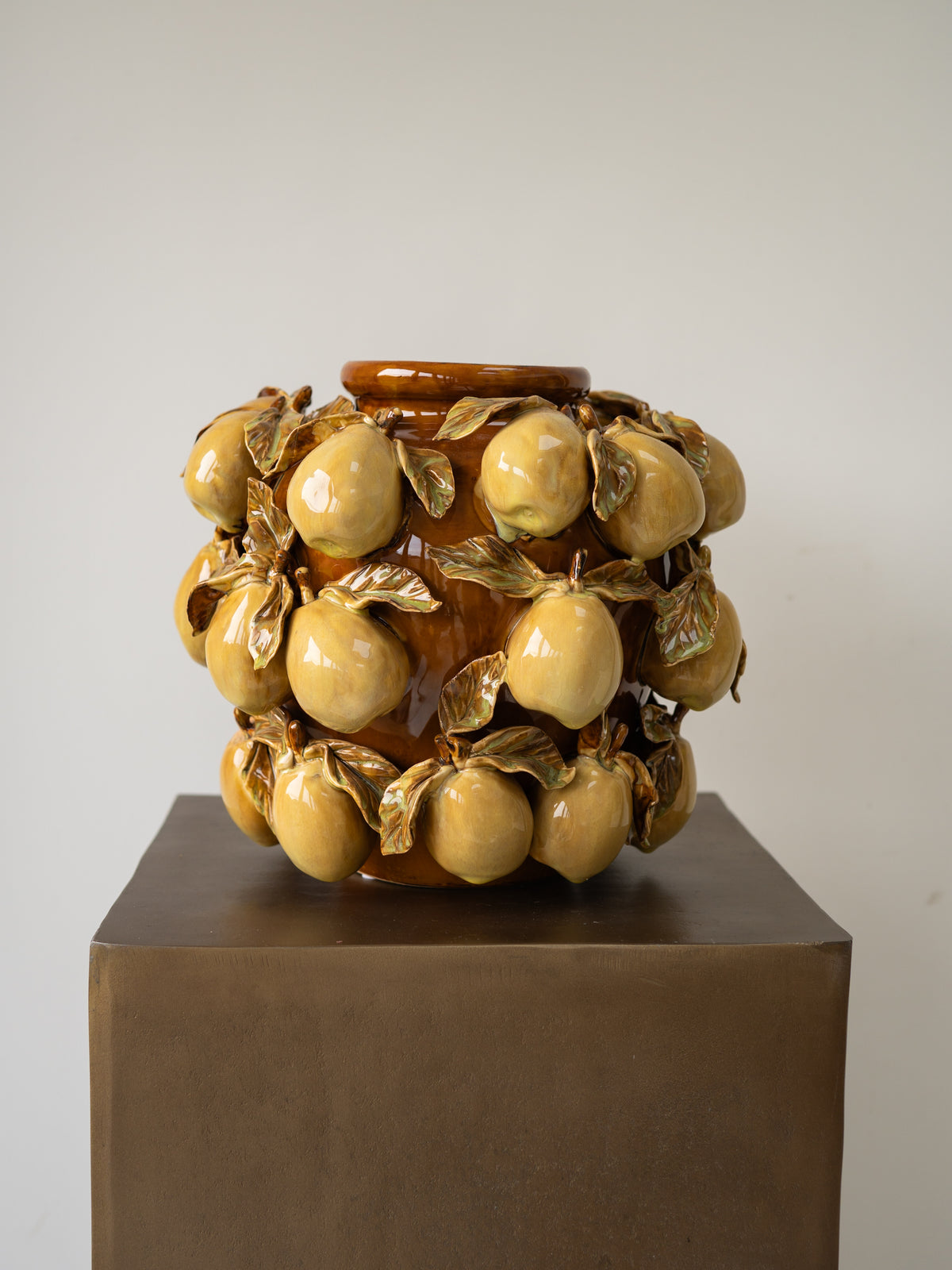 A glazed ceramic sculpture of a vase decorated with numerous glossy yellow-orange pears and metallic gold leaves, displayed on a dark brown pedestal against a neutral light gray background.
