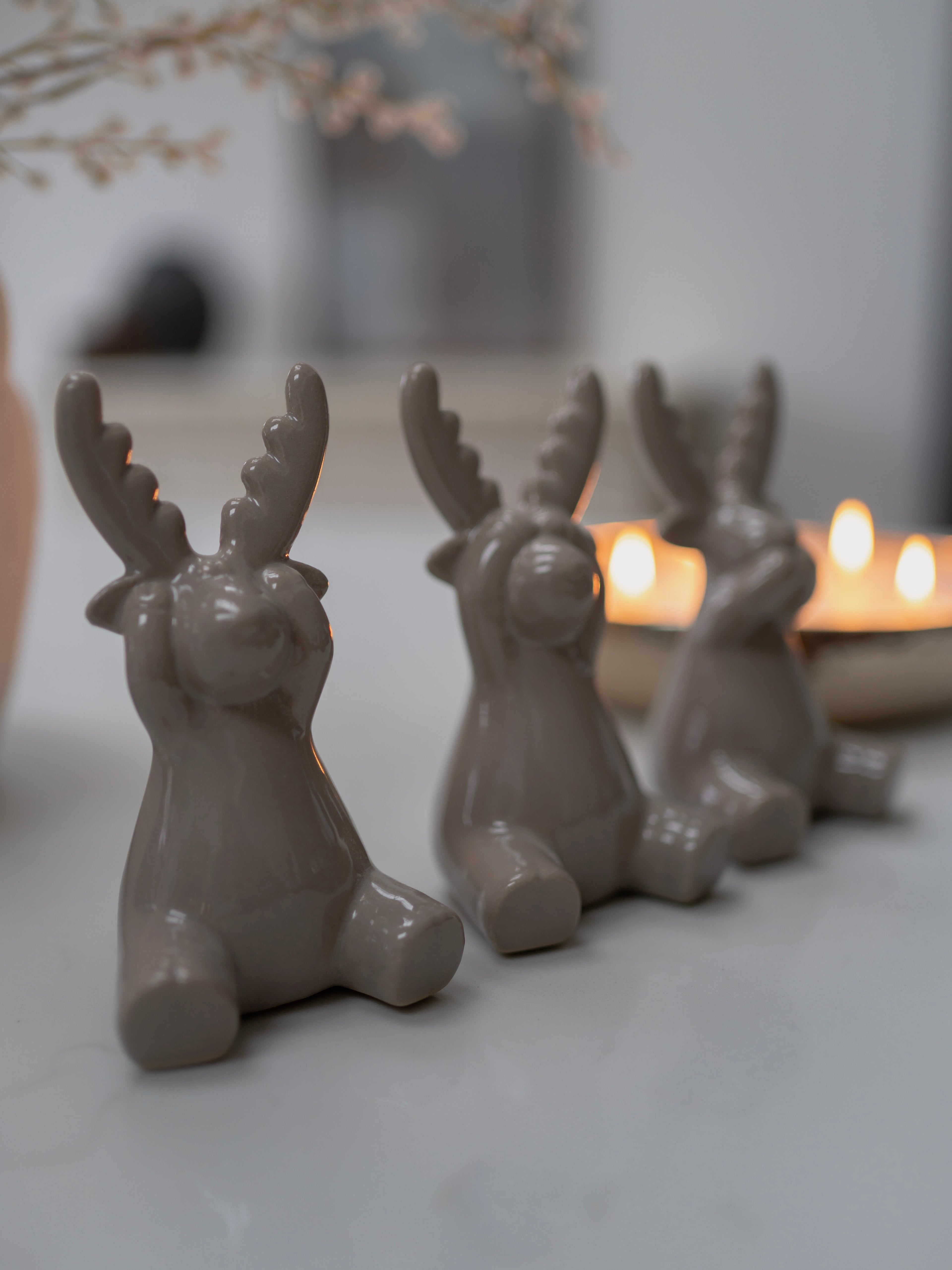 Three grey ceramic deer figurines with antlers on a light surface with candles in the background.