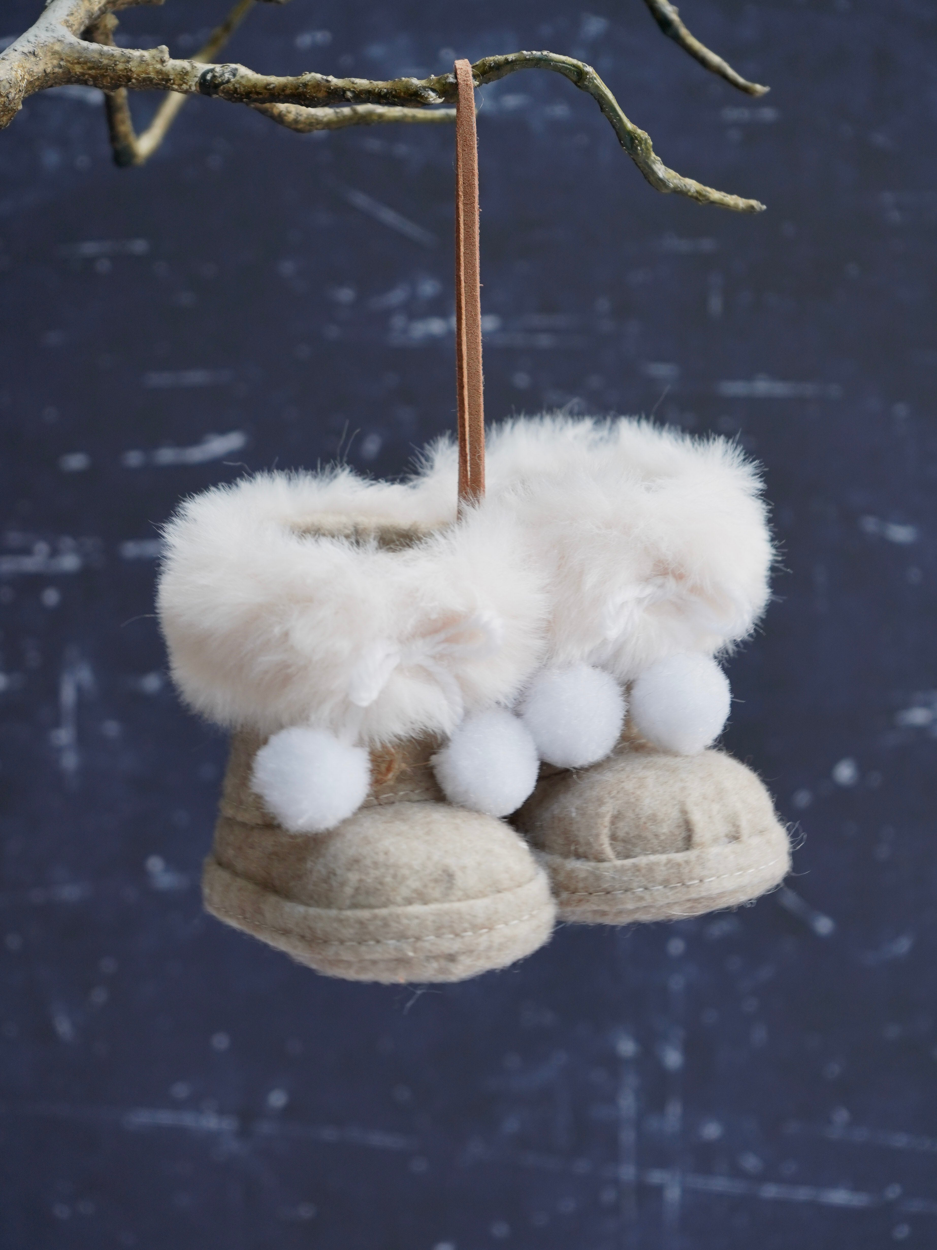Decorative boot ornaments with white fur and pom-poms hanging from a branch against a dark background.