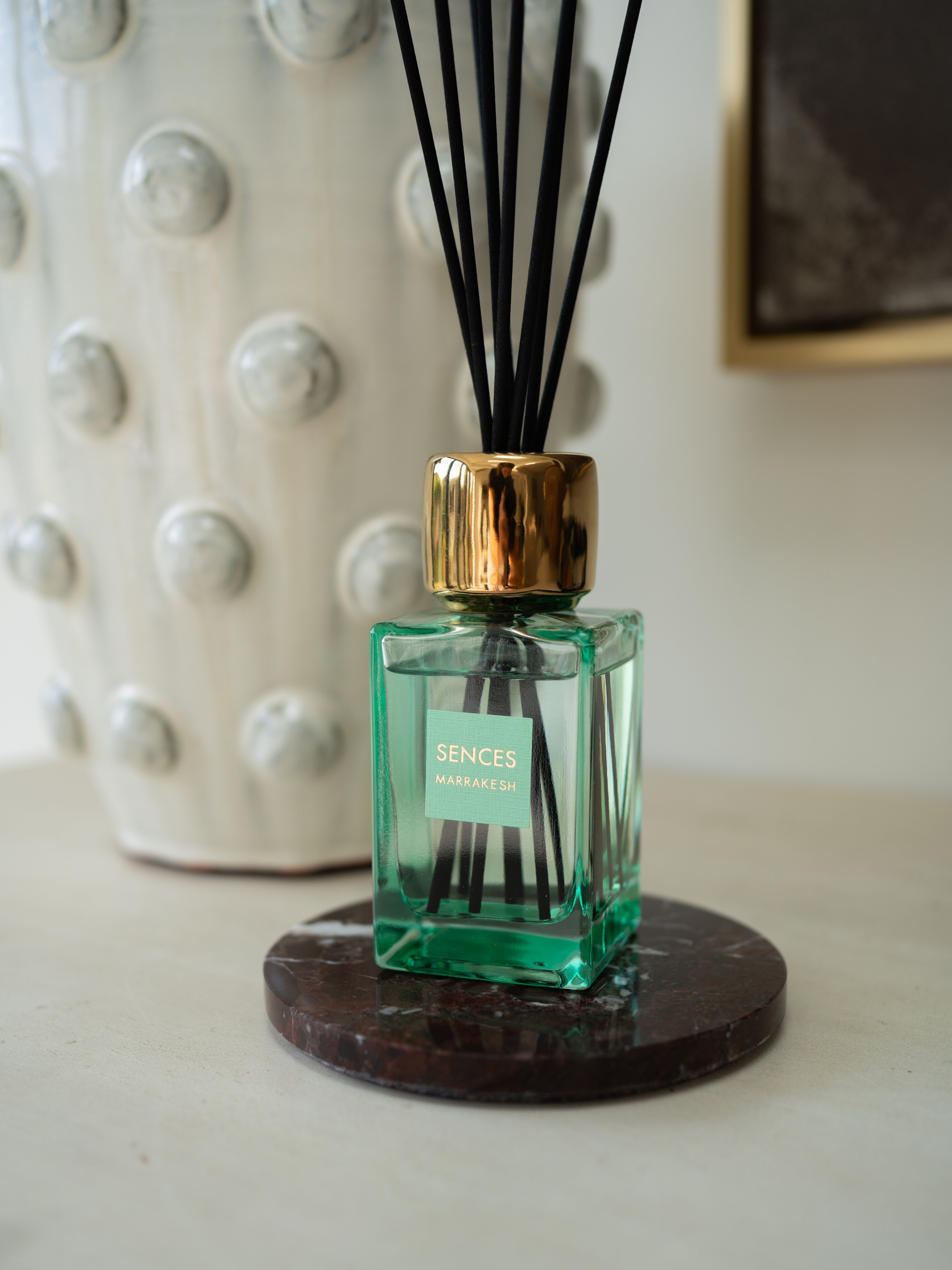 A Senses Marrakesh reed diffuser bottle with a translucent green glass body, a shiny gold cap, and black reeds, resting on a round dark coaster.