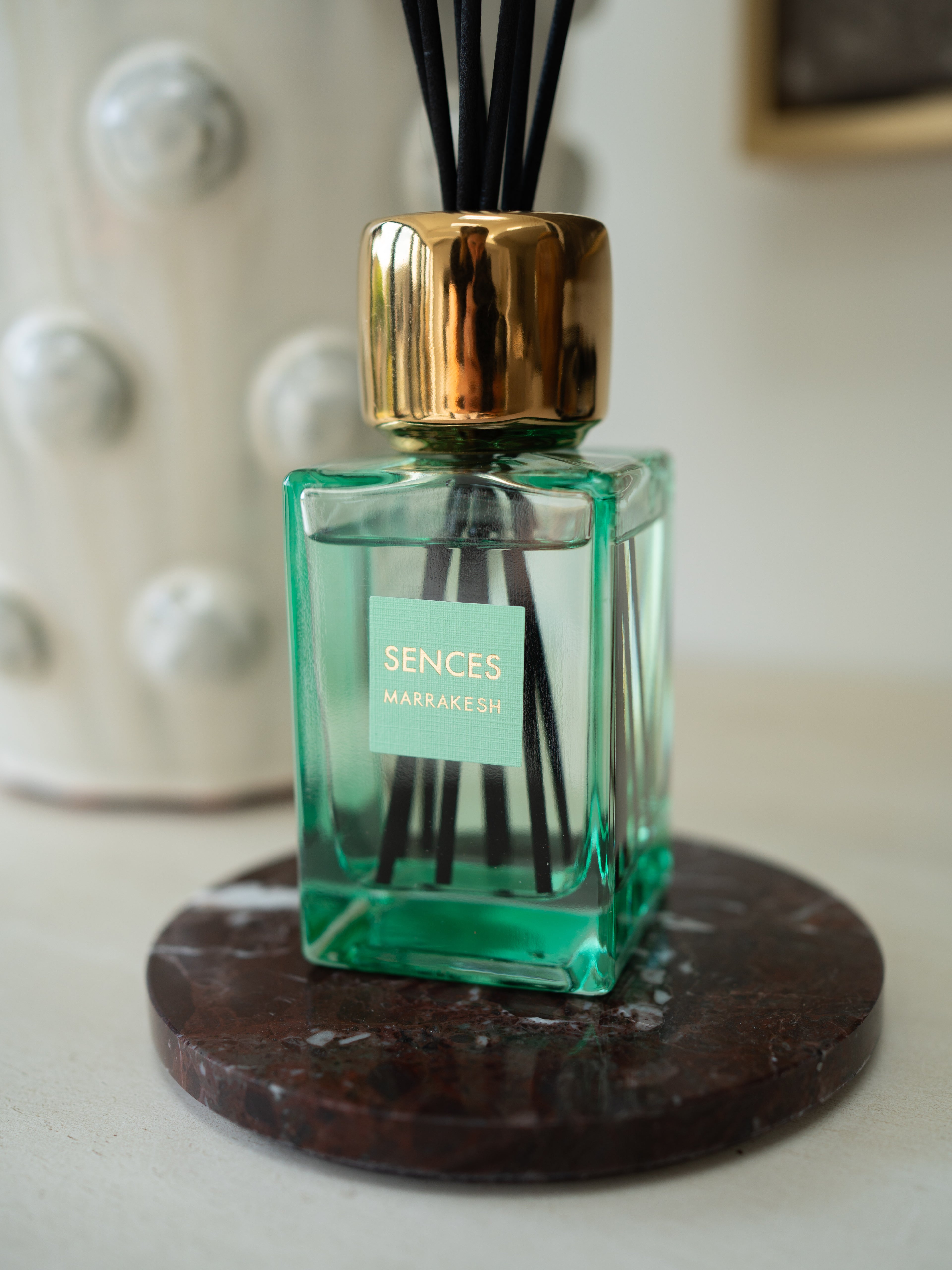 A green glass reed diffuser bottle with a shiny gold cap and black reeds, sitting on a round dark coaster. The label reads &