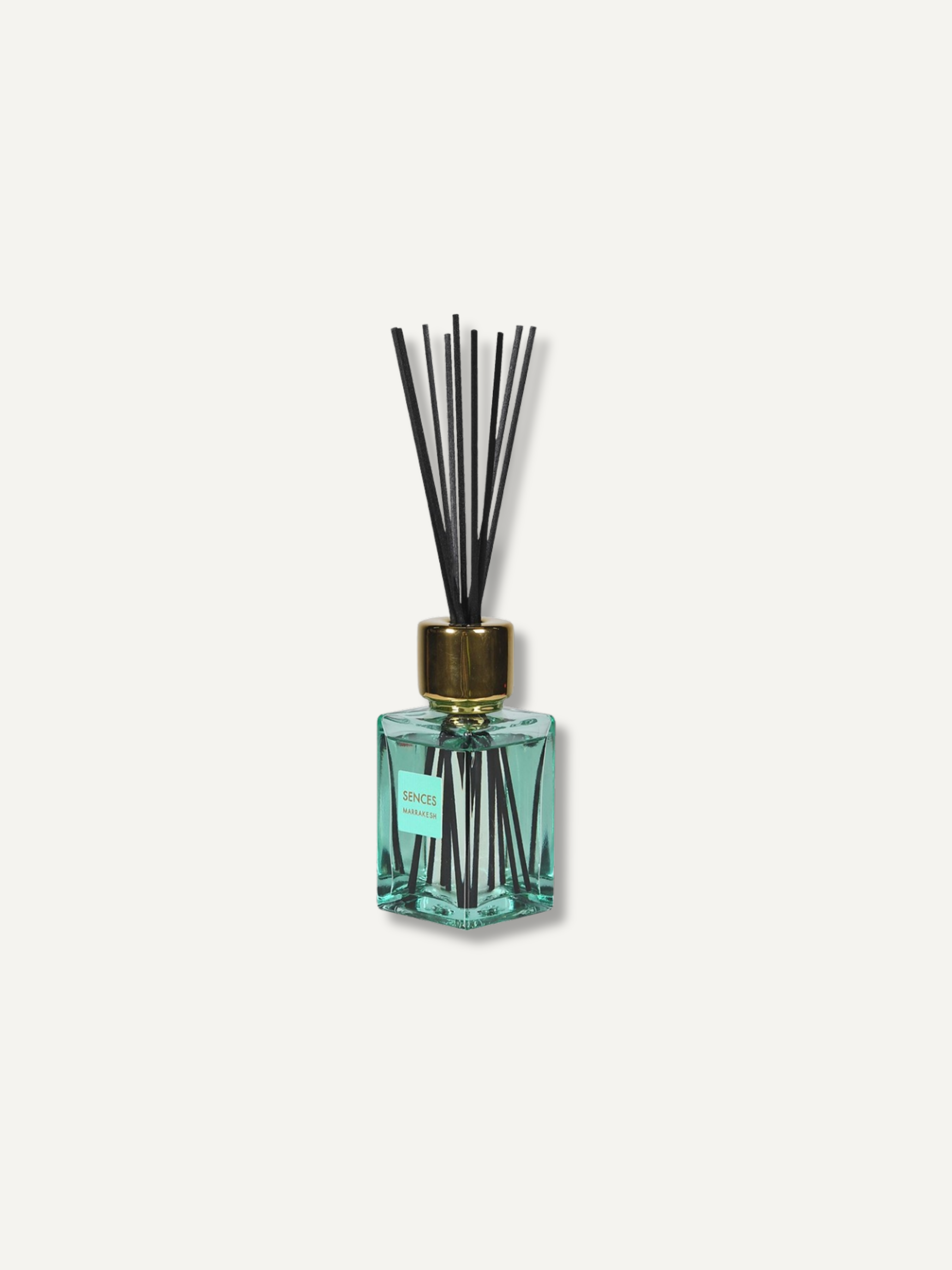 A reed diffuser bottle with a faceted clear glass body filled with aqua liquid, a gold-toned cap, and black reeds, centered on a light warm gray background.