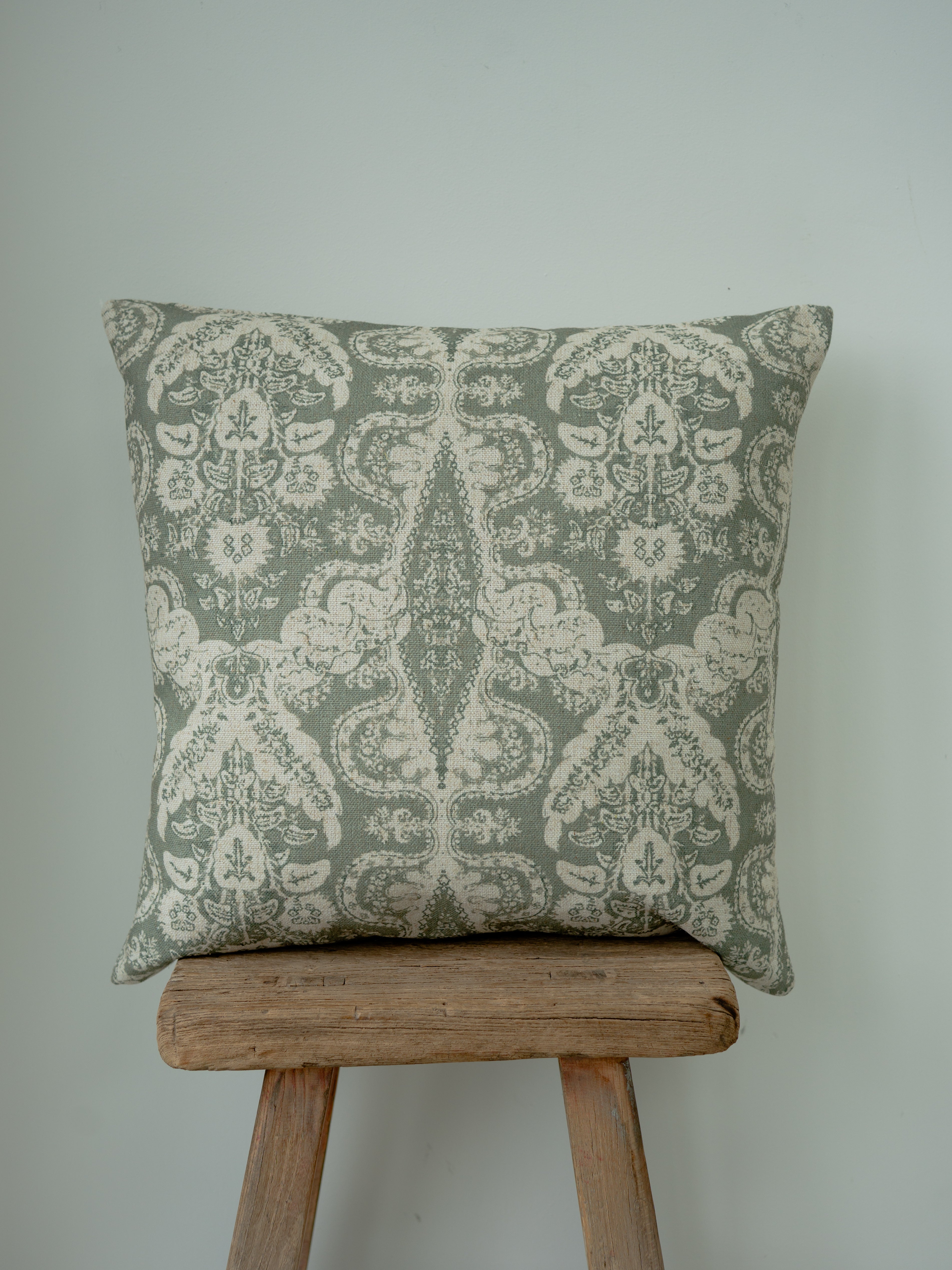 A square throw cushion with a muted sage green and cream damask pattern, resting on a rustic wooden stool against a plain light gray wall.