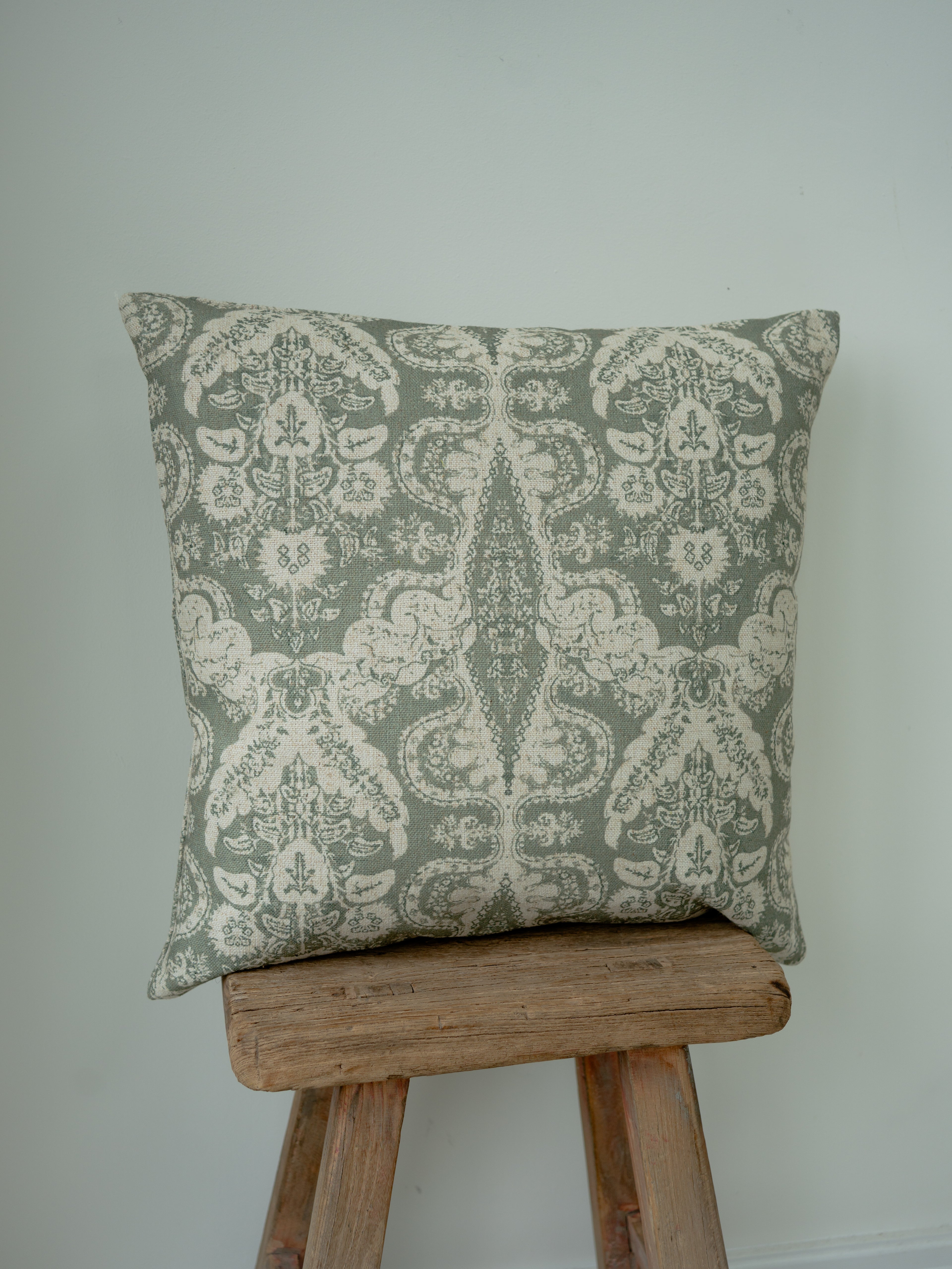 A square decorative throw cushion with a muted sage green and cream damask pattern, displayed on a rustic wooden stool against a plain light gray wall.