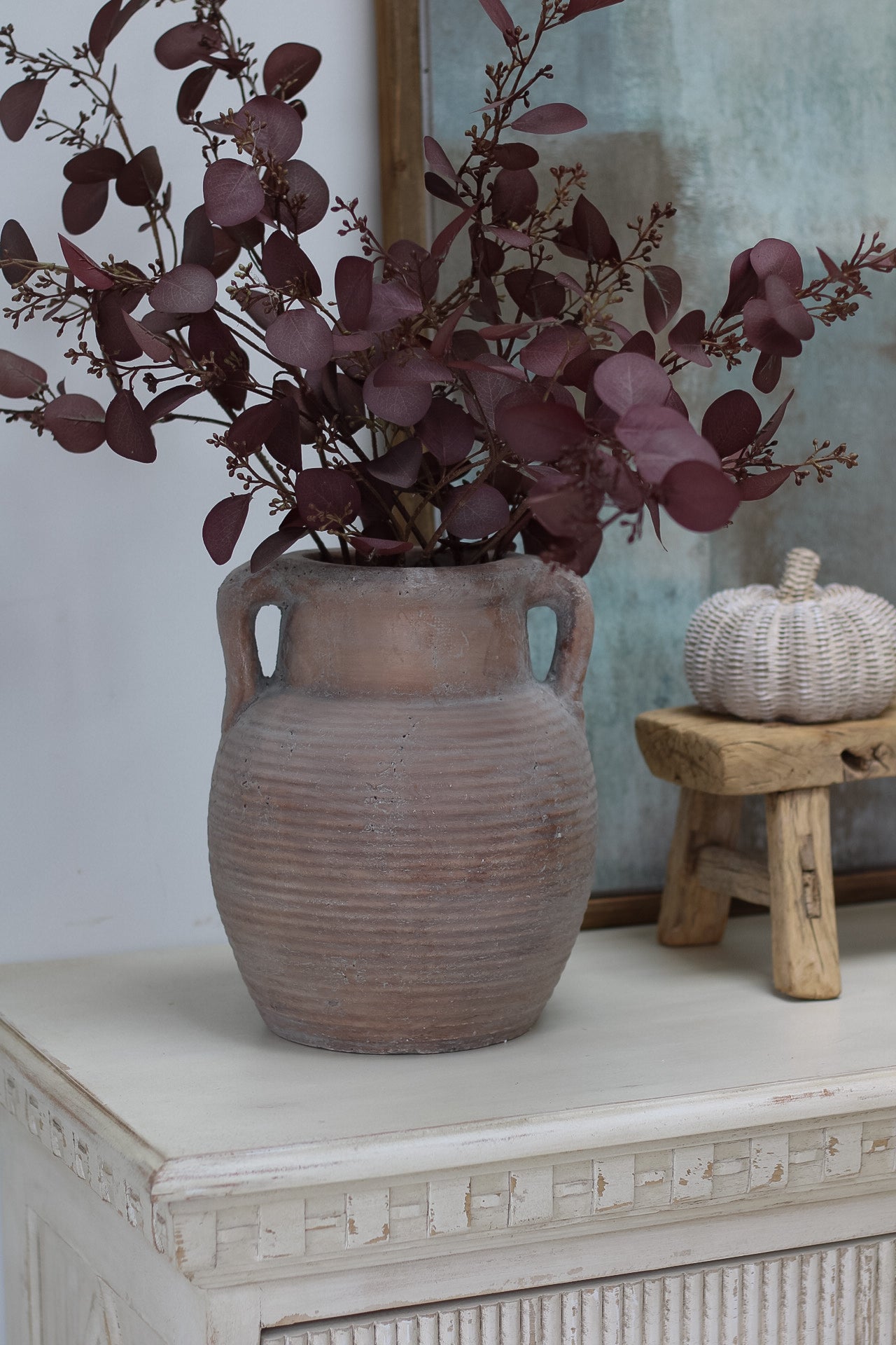 Decorative vase with purple foliage on a white surface with a wooden stool and pumpkin in the background.