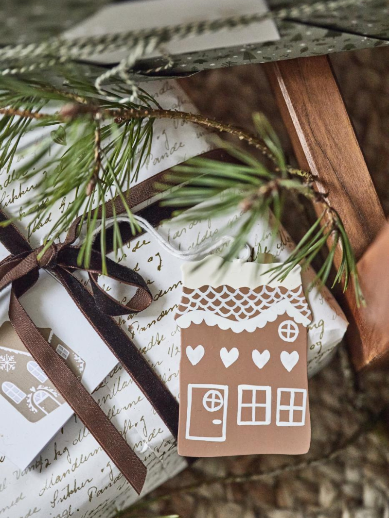 Gift wrapped in white paper with a brown house-shaped tag and pine branch, on a wooden surface.