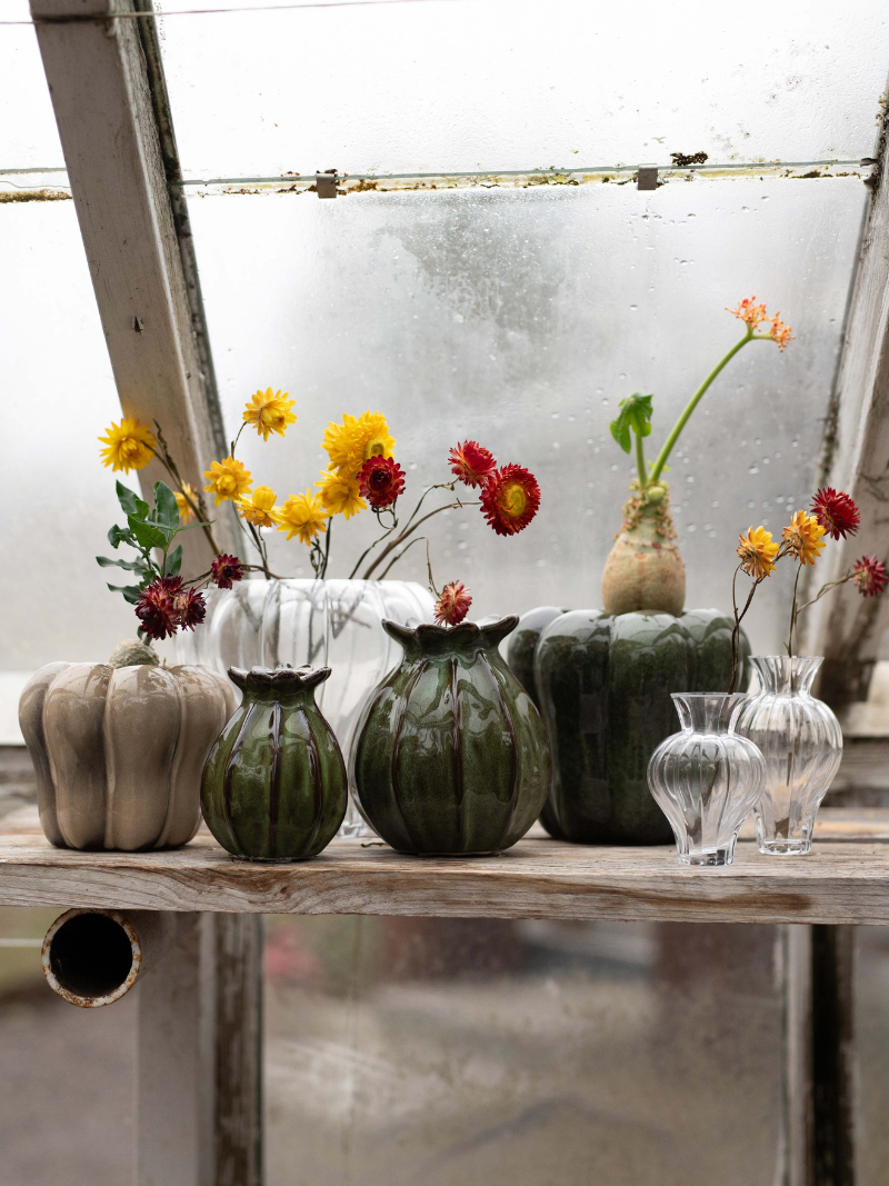Decorative poppy head vases and flower arrangements on a wooden table with a glass paneled wall in the background.