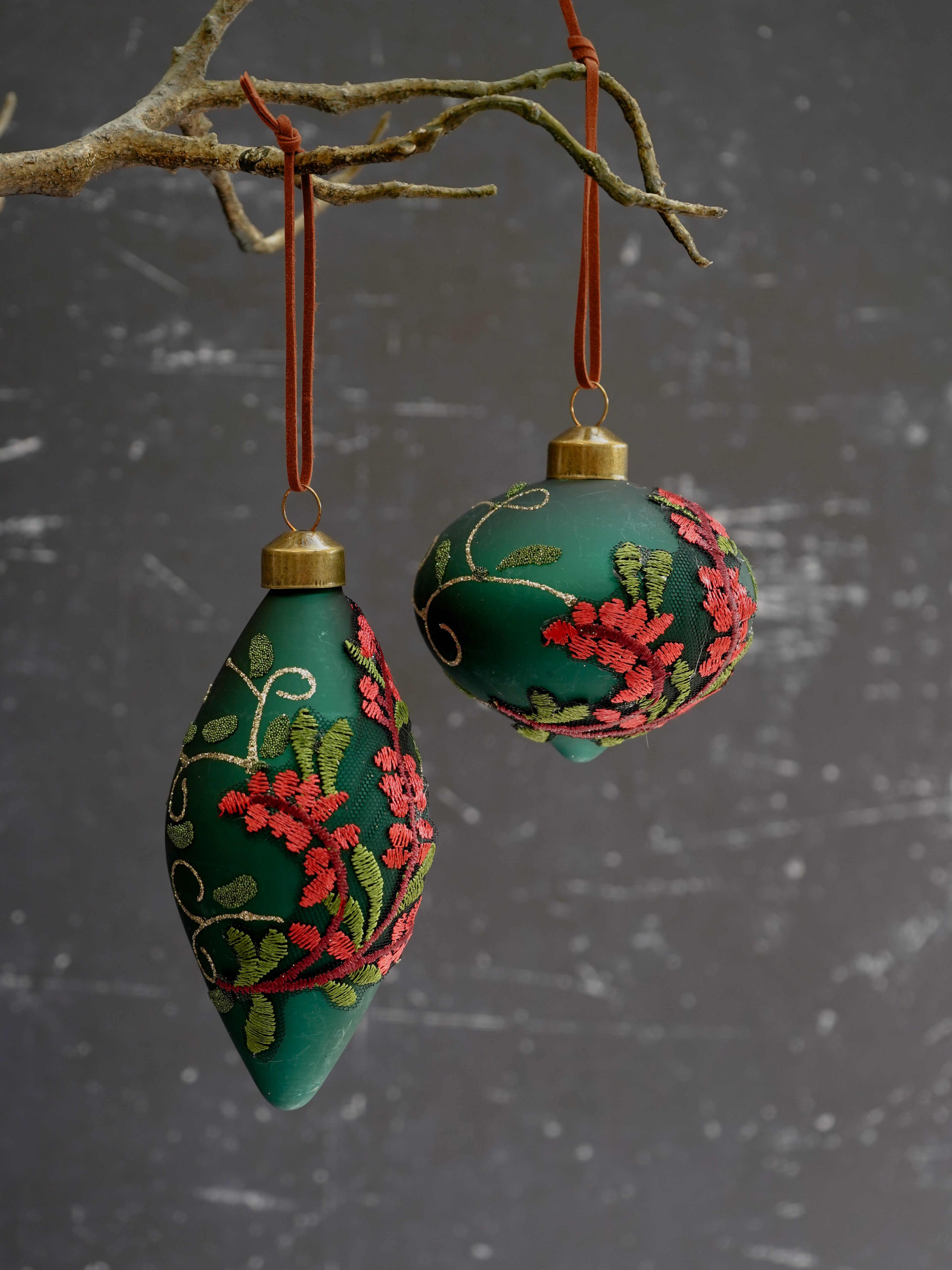 Two green embroidered ornaments hanging from branches against a gray background