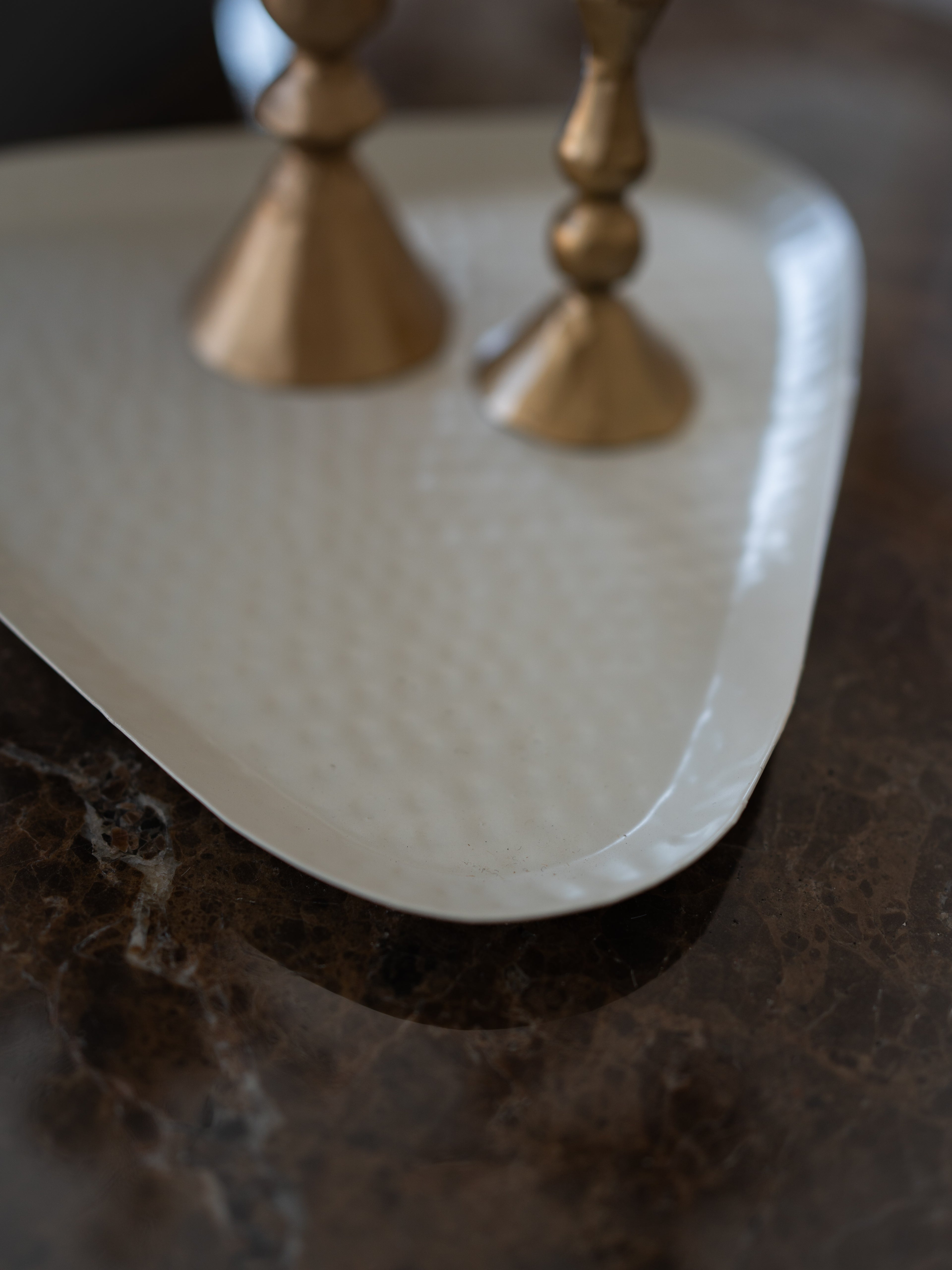 A close-up of a white hammered metal serving tray with curved edges, resting on a dark brown marble surface with two gold-toned candlesticks partially visible in the background.