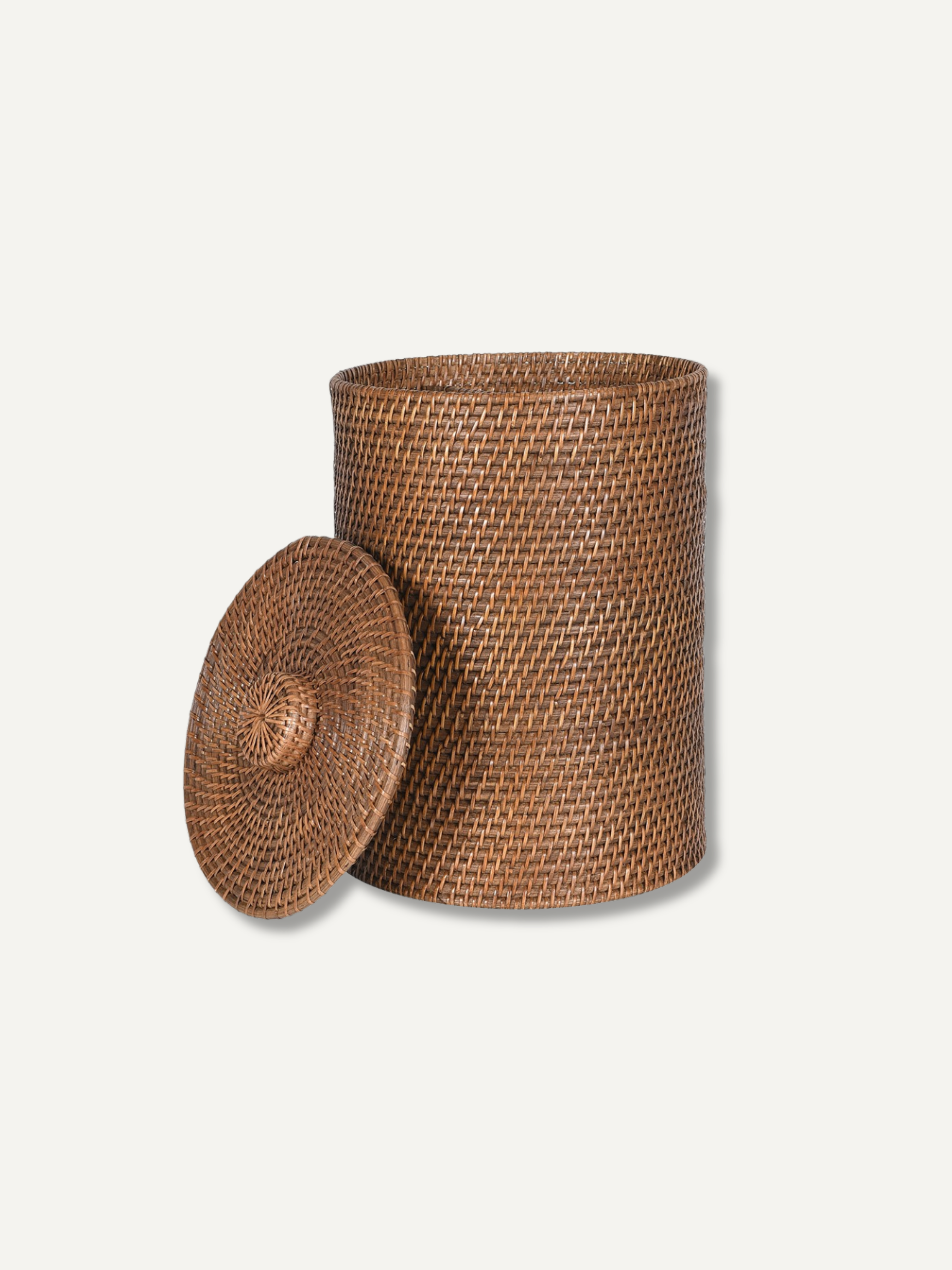 A natural brown woven rattan storage basket with a matching round lid leaning against it, set against a plain off-white background.