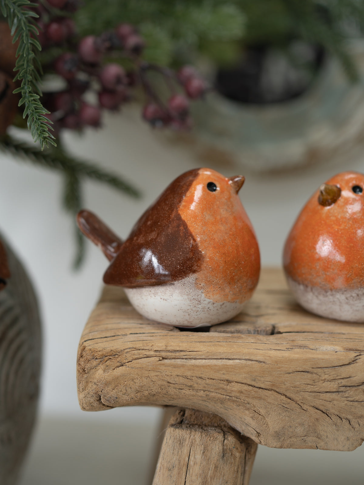 Two small glazed robin bird figurines in orange, brown, and off-white ceramic, displayed on a rustic wooden stand with a softly blurred holiday background.