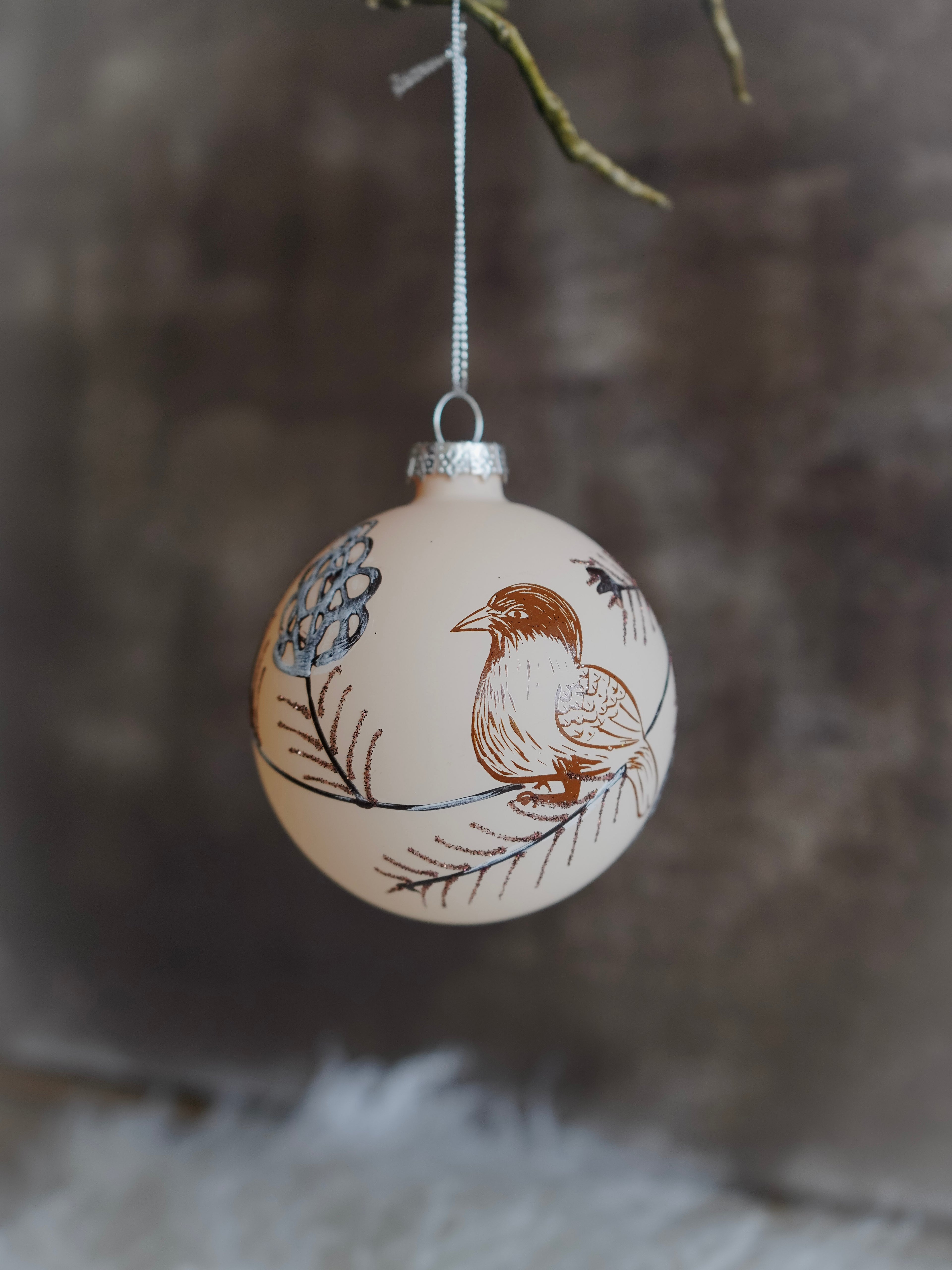 Decorative Christmas ball ornament with bird and floral design hanging against a blurred natural background.