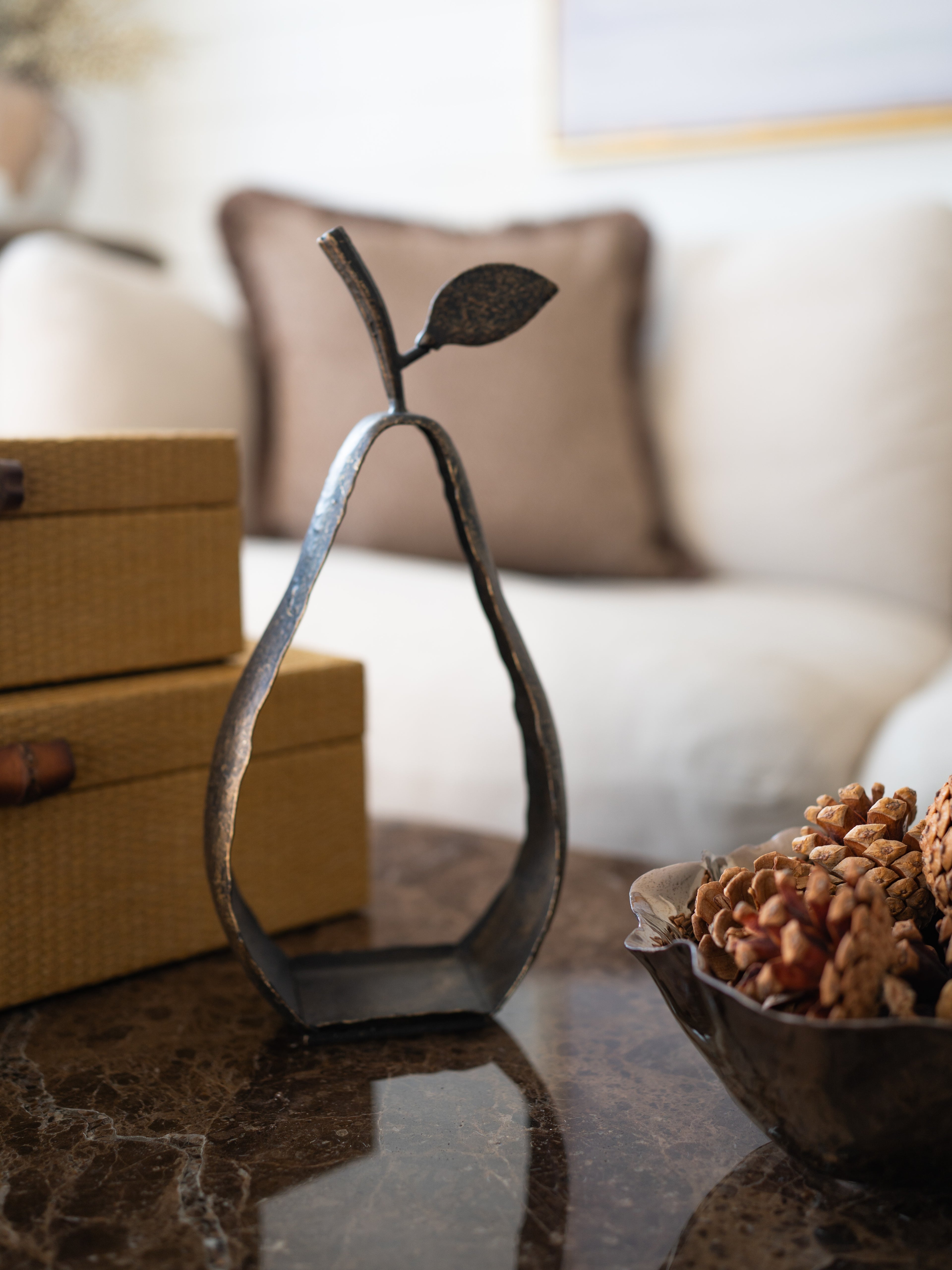 A dark metal decorative sculpture shaped like a stylized pear with a leafy stem, displayed on a polished dark marble table in a living room setting next to a bowl of pinecones and stacked woven boxes.