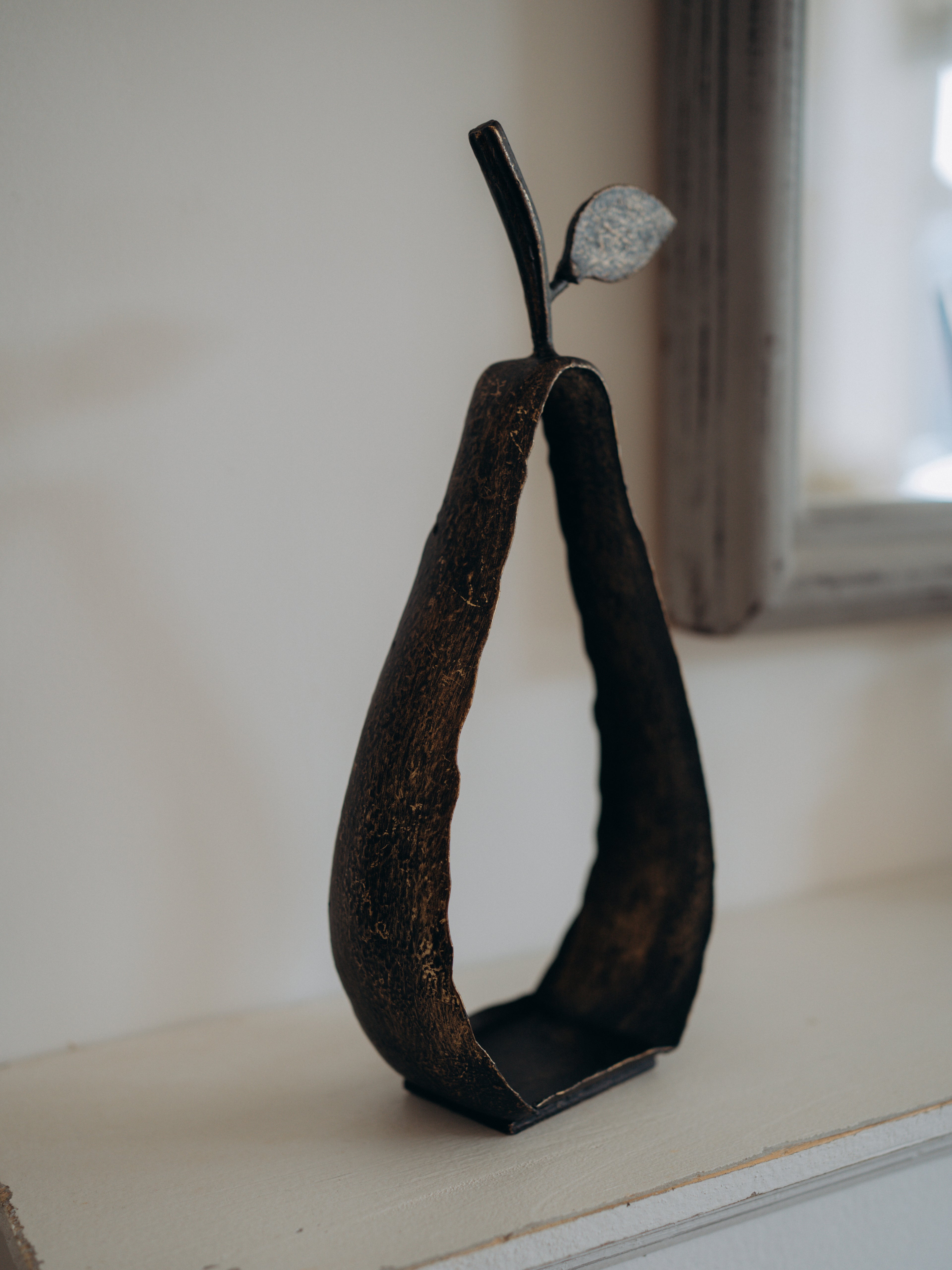 A dark metal decorative sculpture shaped like a pear with a textured, rustic finish and a small leaf on top, displayed on a light-colored windowsill.