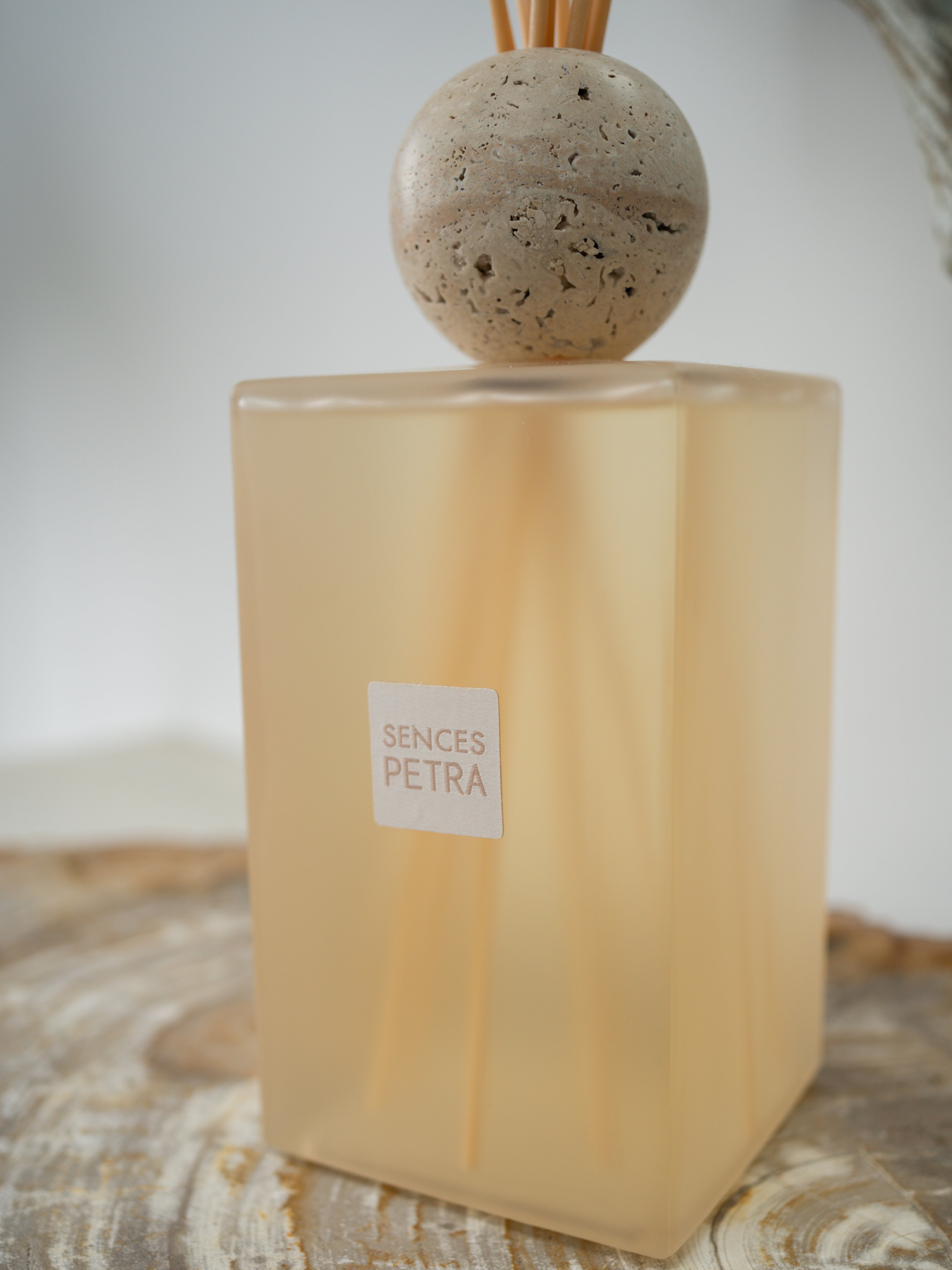 A Sences Petra reed diffuser with a translucent frosted beige glass bottle, natural fossilled wood diffuser sticks, and a round stone top, resting on a rustic wooden surface.