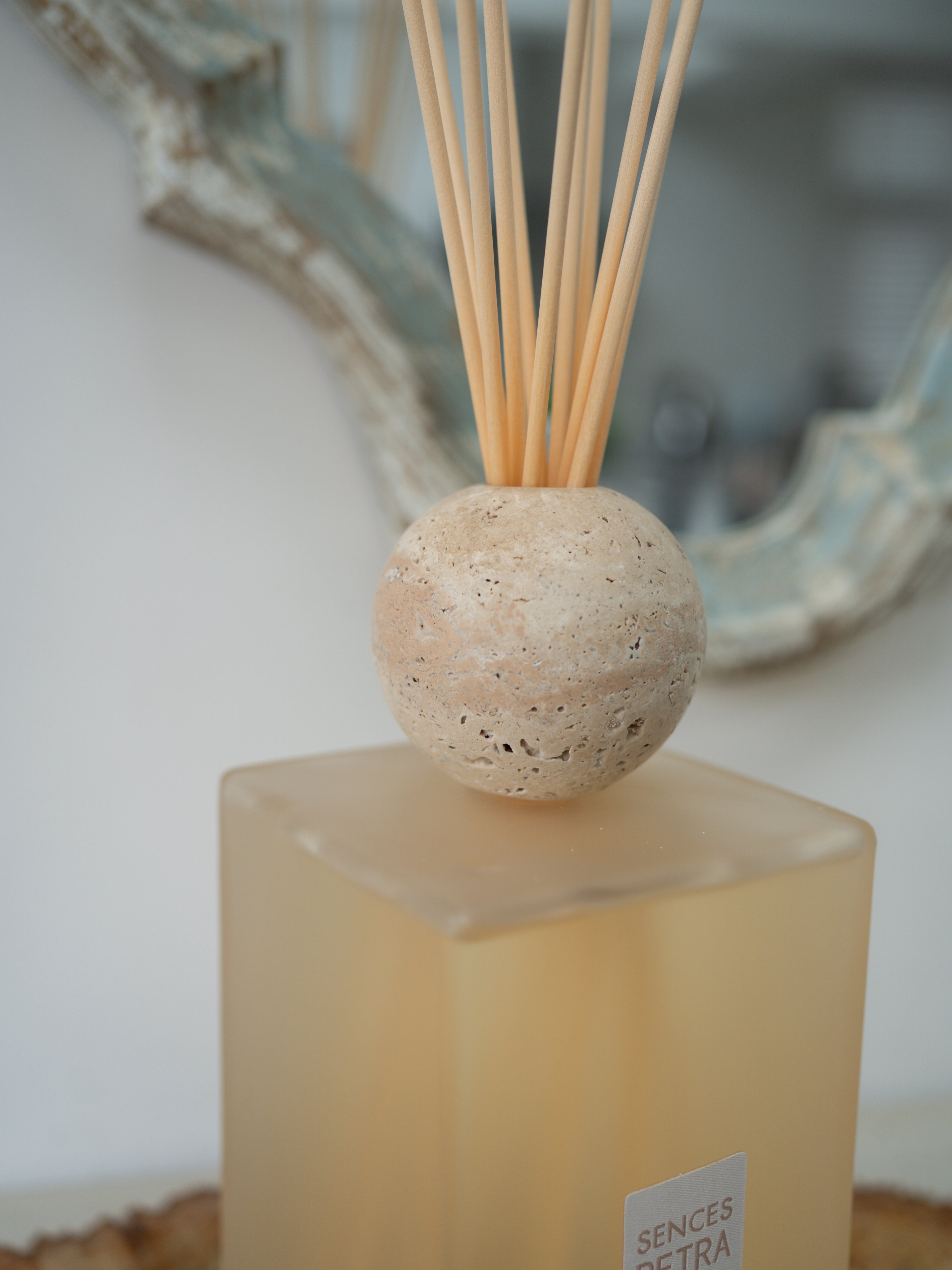 A reed diffuser with a round beige travertine stone sphere on a frosted amber glass base, with natural reed sticks inserted, photographed indoors against a softly blurred background.