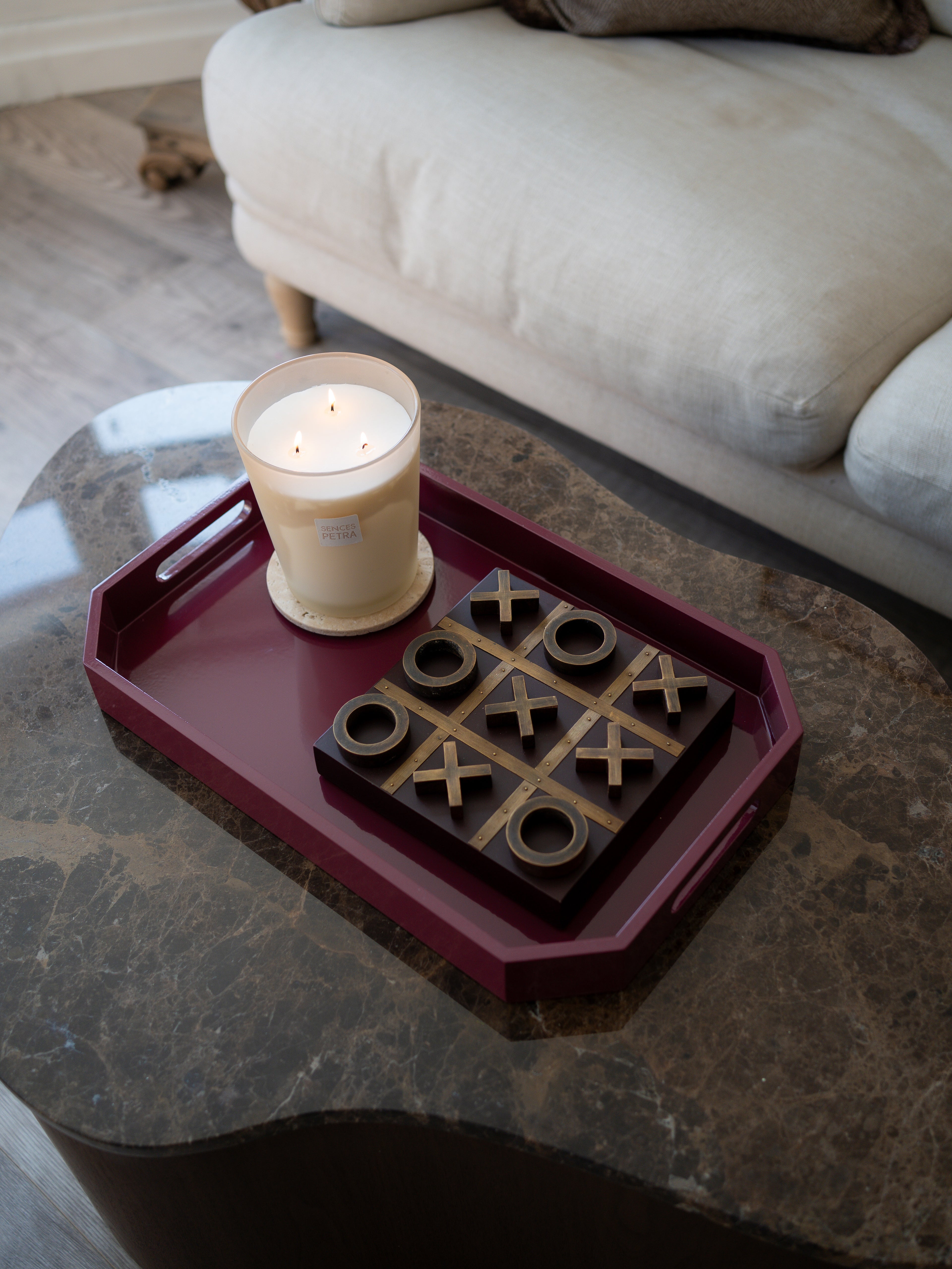 A dark burgundy rectangular serving tray on a dark marble coffee table, holding a black and gold wooden tic-tac-toe game and a lit cream-colored candle. A light-colored sofa is visible in the background.