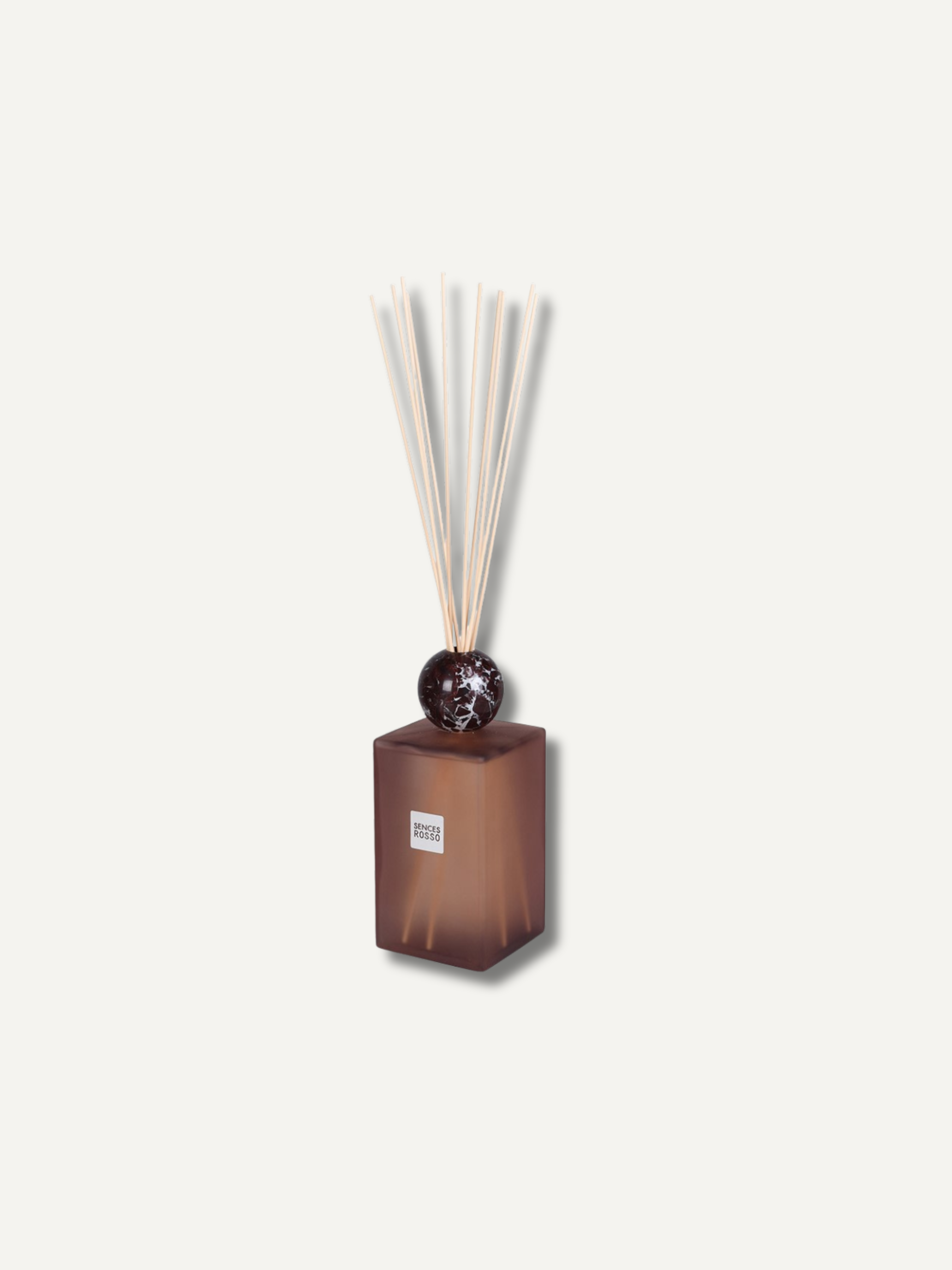 A reed diffuser with a translucent amber-brown glass bottle, a dark round stopper, and natural reed sticks, shown against a solid off-white background.