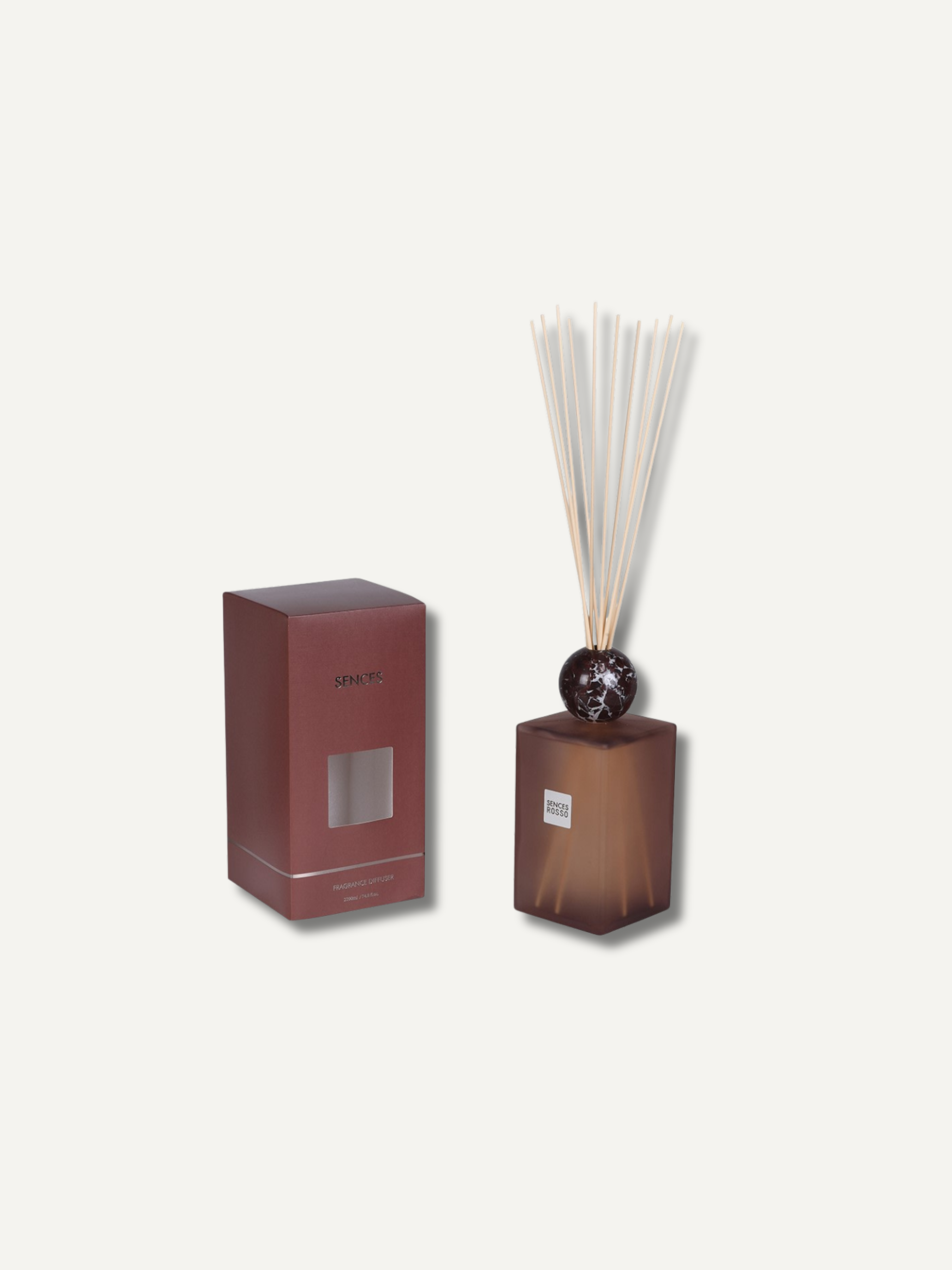 A reed diffuser set with a translucent amber-brown glass bottle, dark round stopper, and natural reeds, shown next to its matching rectangular box on a light cream background.