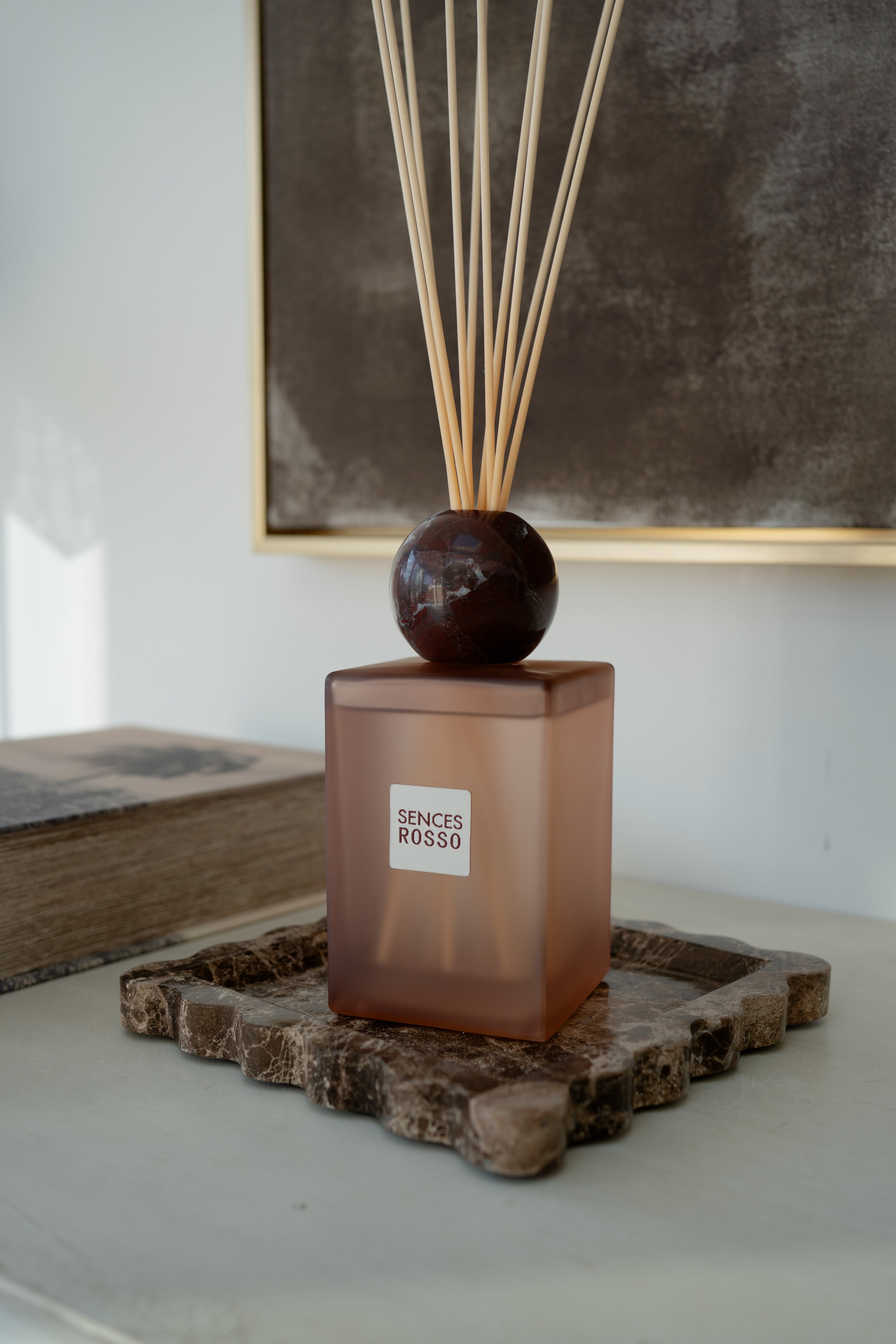 A reed diffuser bottle with a frosted amber-brown glass body, a dark round stopper, and natural reed sticks, displayed on a rustic stone tray in an indoor setting.