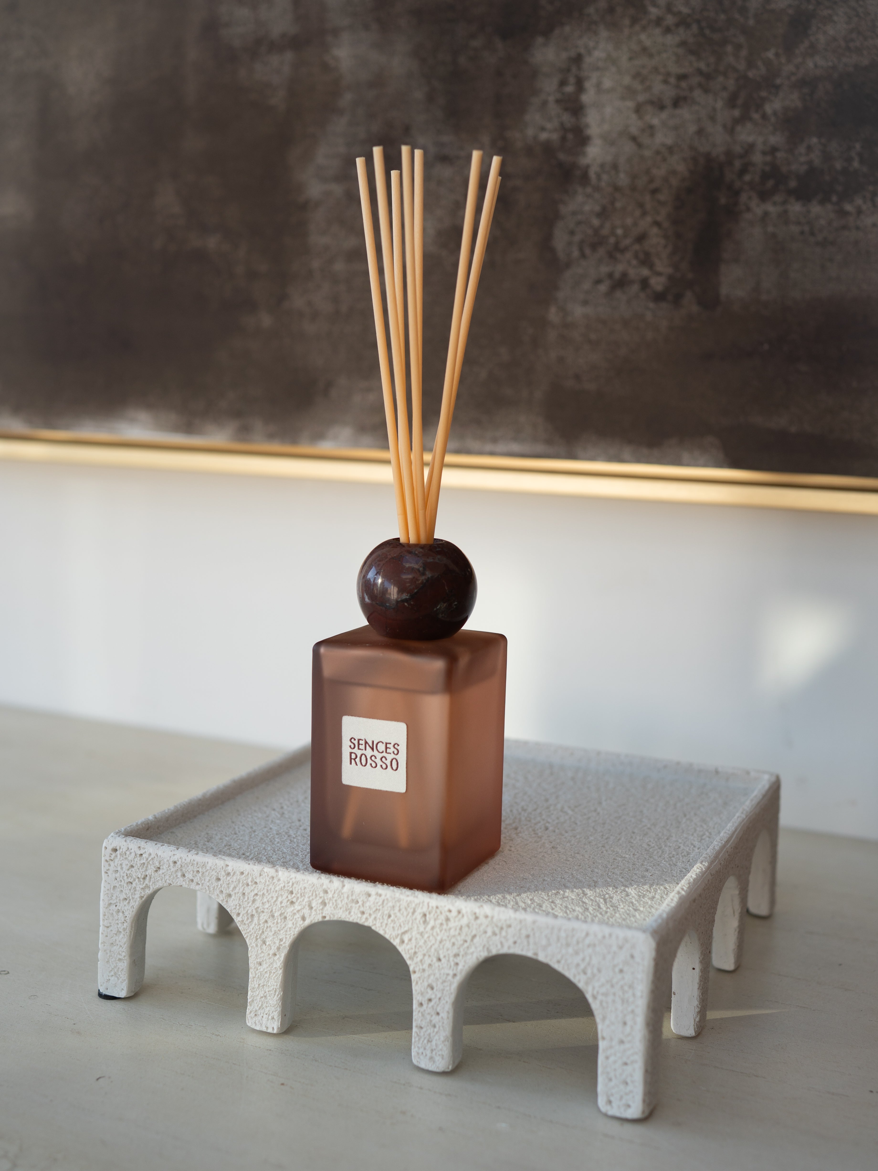 A reed diffuser bottle with a frosted amber-brown glass body, a dark round stopper, and natural reed sticks, displayed on a white textured pedestal against a neutral indoor background.