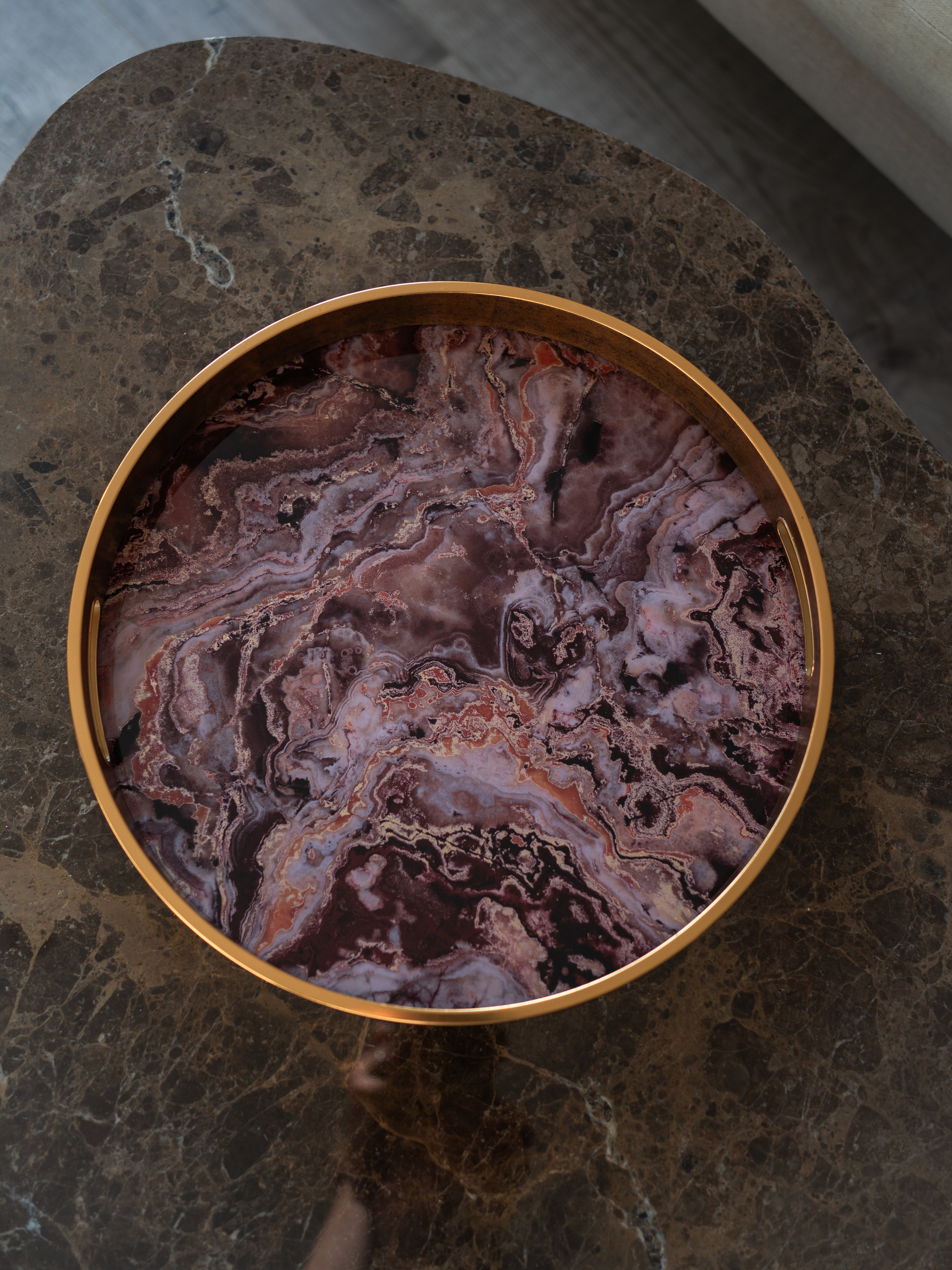 A round decorative tray with a polished gold-toned rim and a marbled purple, pink, and brown agate-like pattern, shown from above on a dark stone surface.
