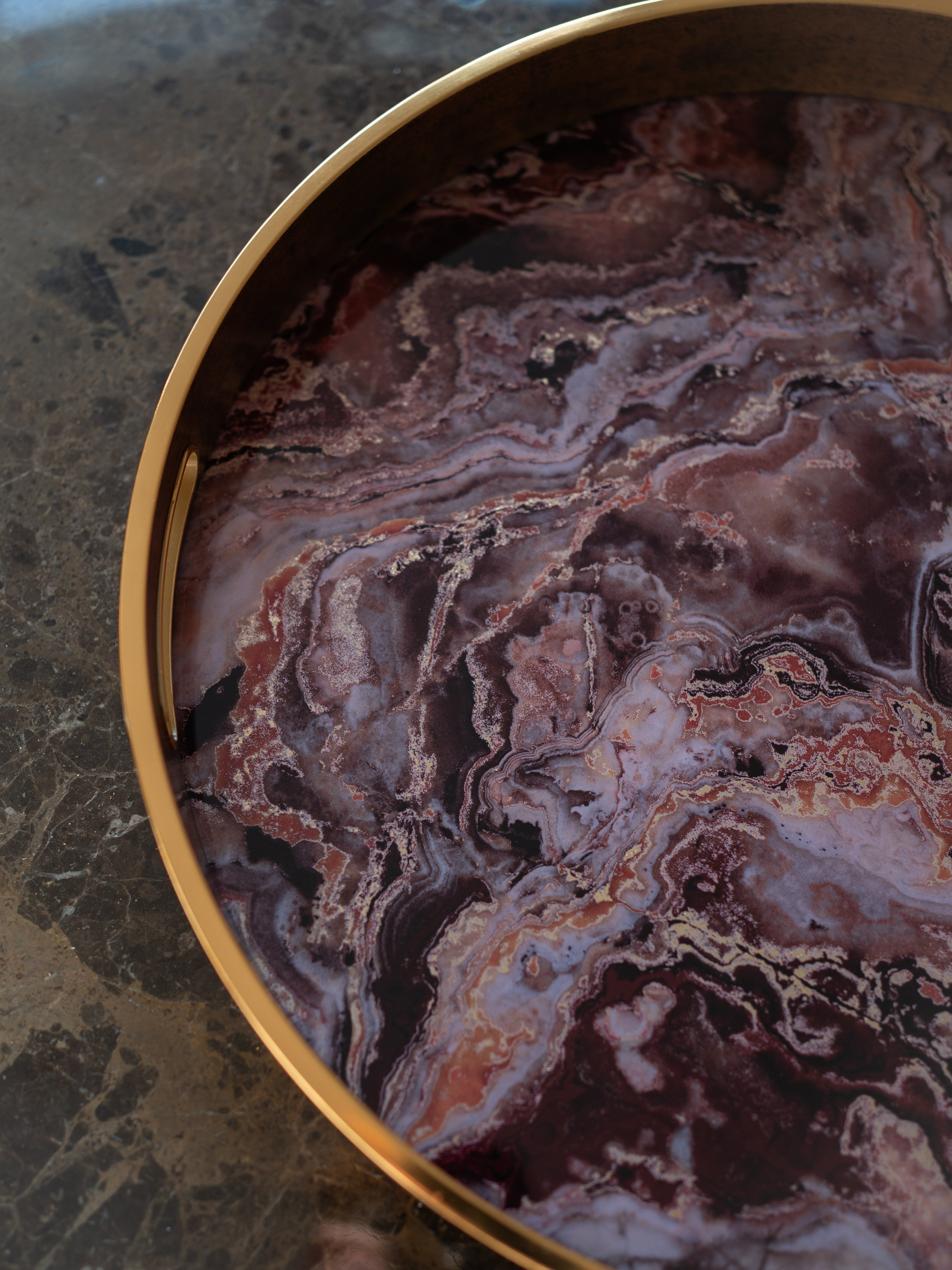 A close-up of a round decorative tray with a polished gold-toned rim and a marbled stone insert featuring swirling patterns of purple, burgundy, brown, and cream, set on a dark marble surface.