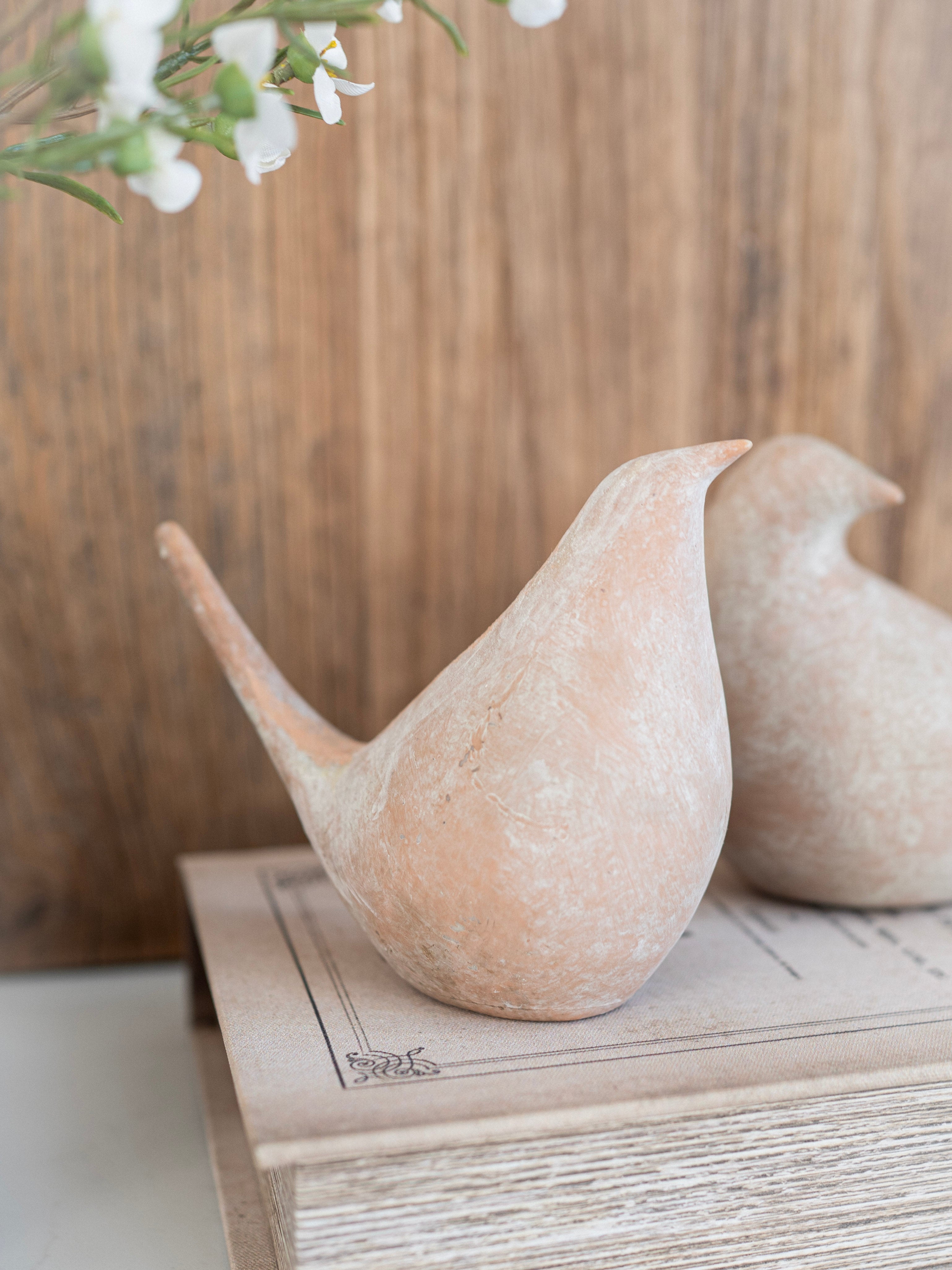Two beige bird-shaped sculptures on a wooden surface with a blurred floral background