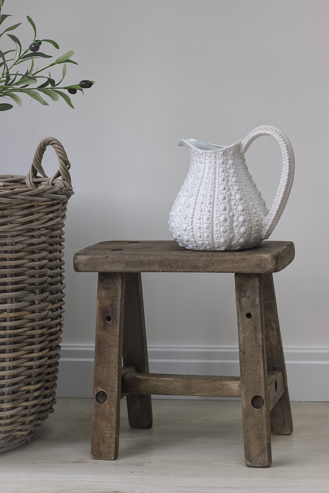 A rustic wooden stool made from pine wood, displayed in a room setting with a white ceramic jug on top.