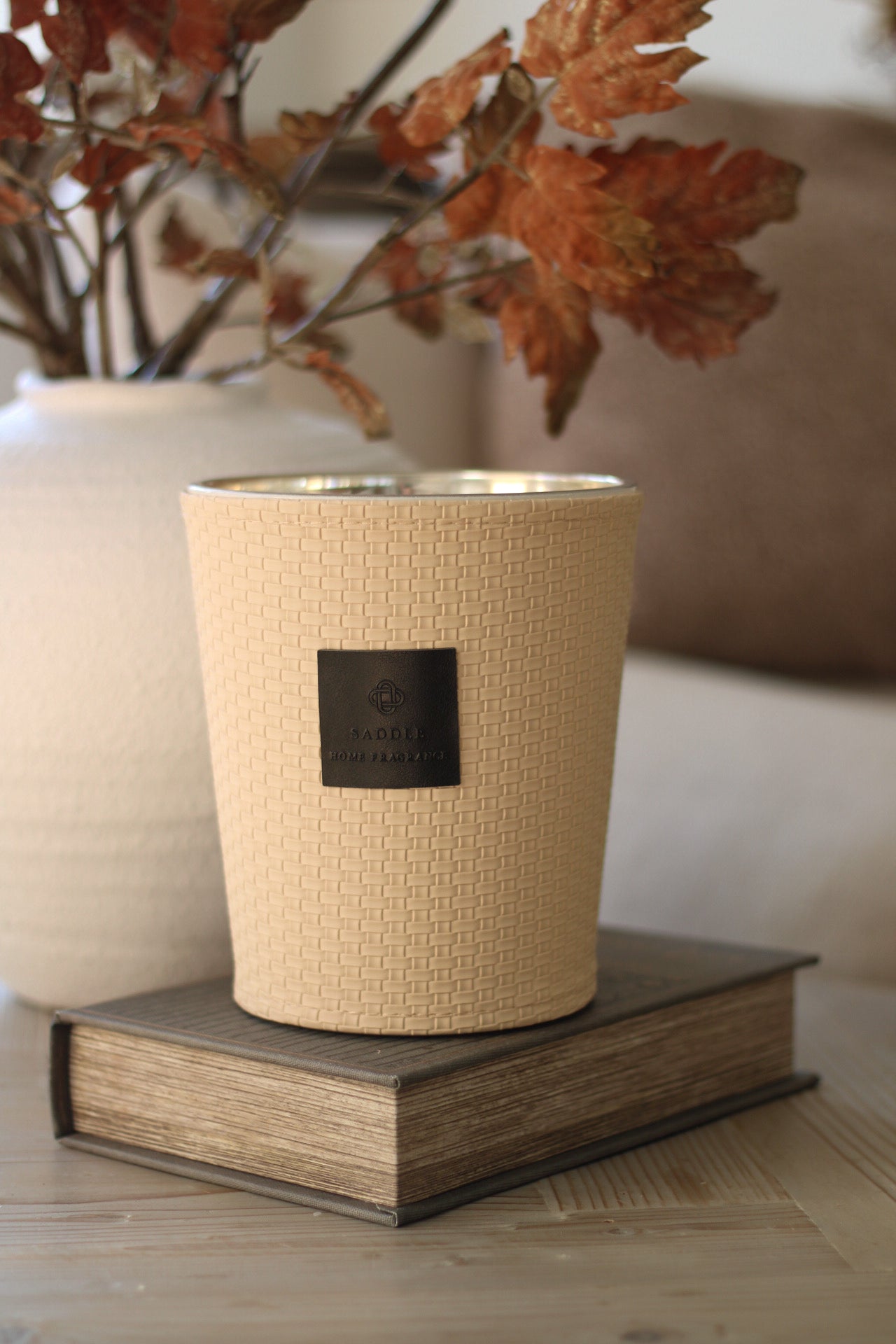 Beige textured candle on a book with a vase of dried leaves in the background