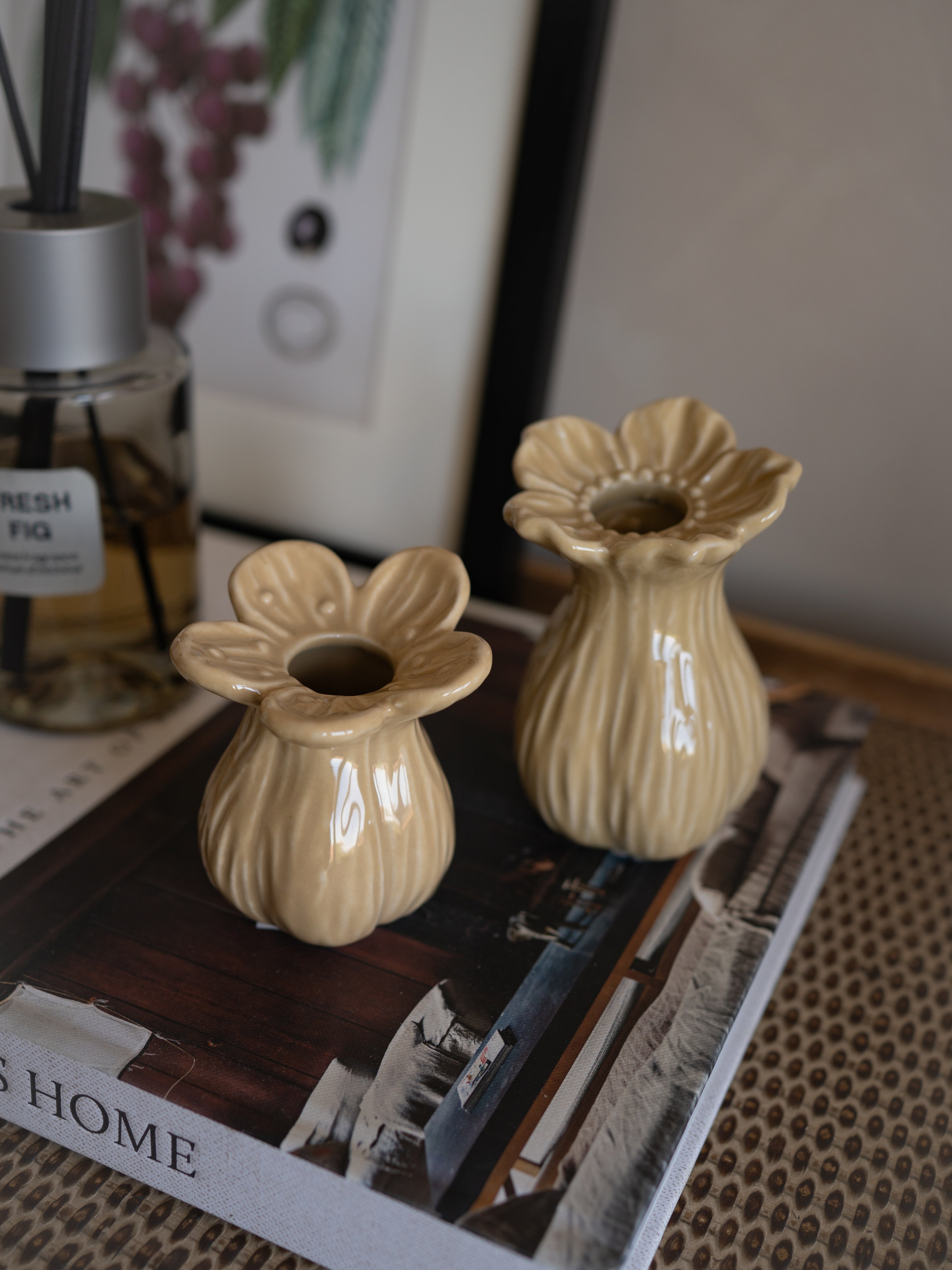 Two beige ceramic flower-shaped bud vases on a magazine with a diffuser and plant in the background.