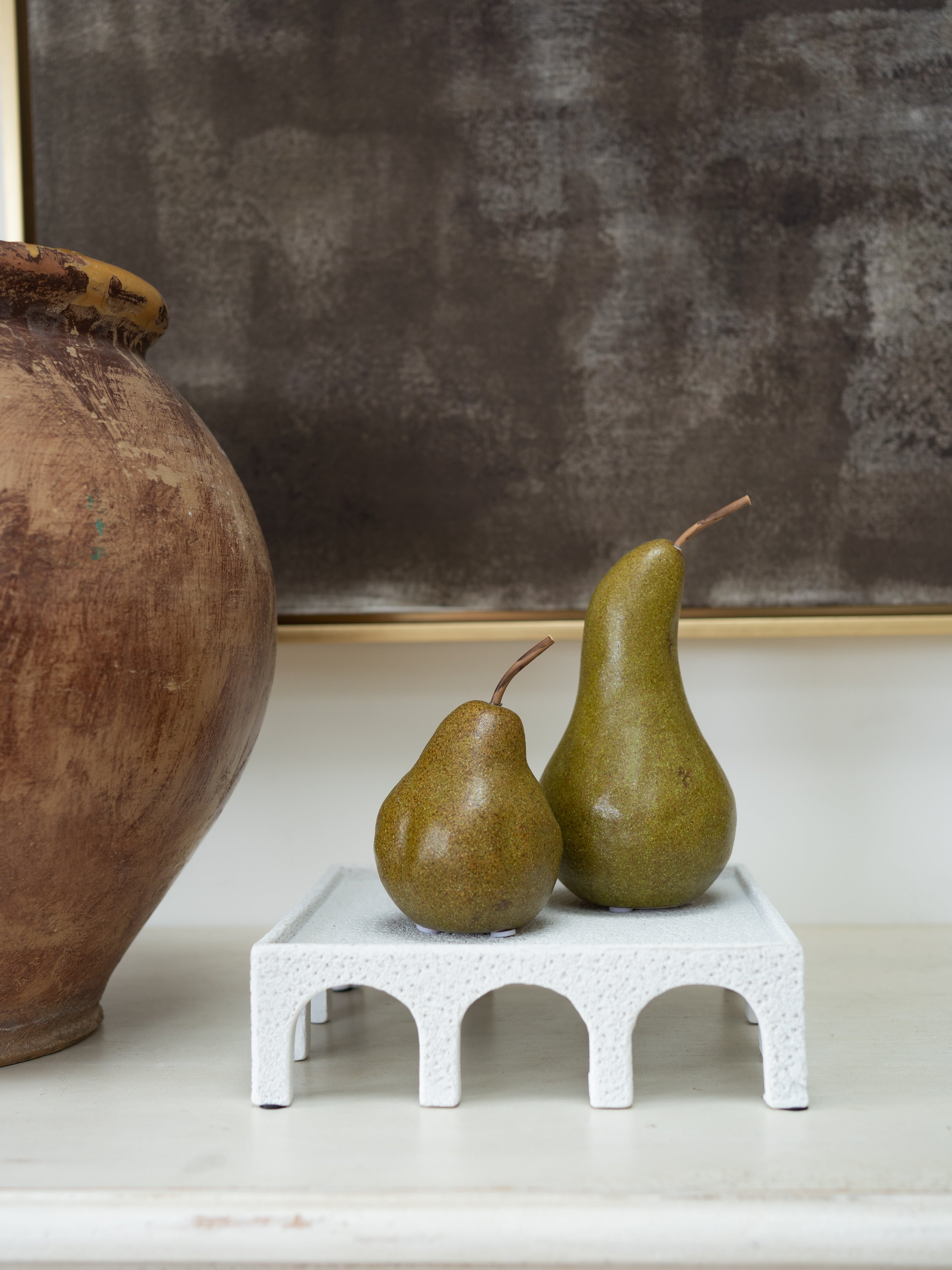 Two pears on a small white arched stand, with a large brown ceramic vase and a dark textured wall in the background.