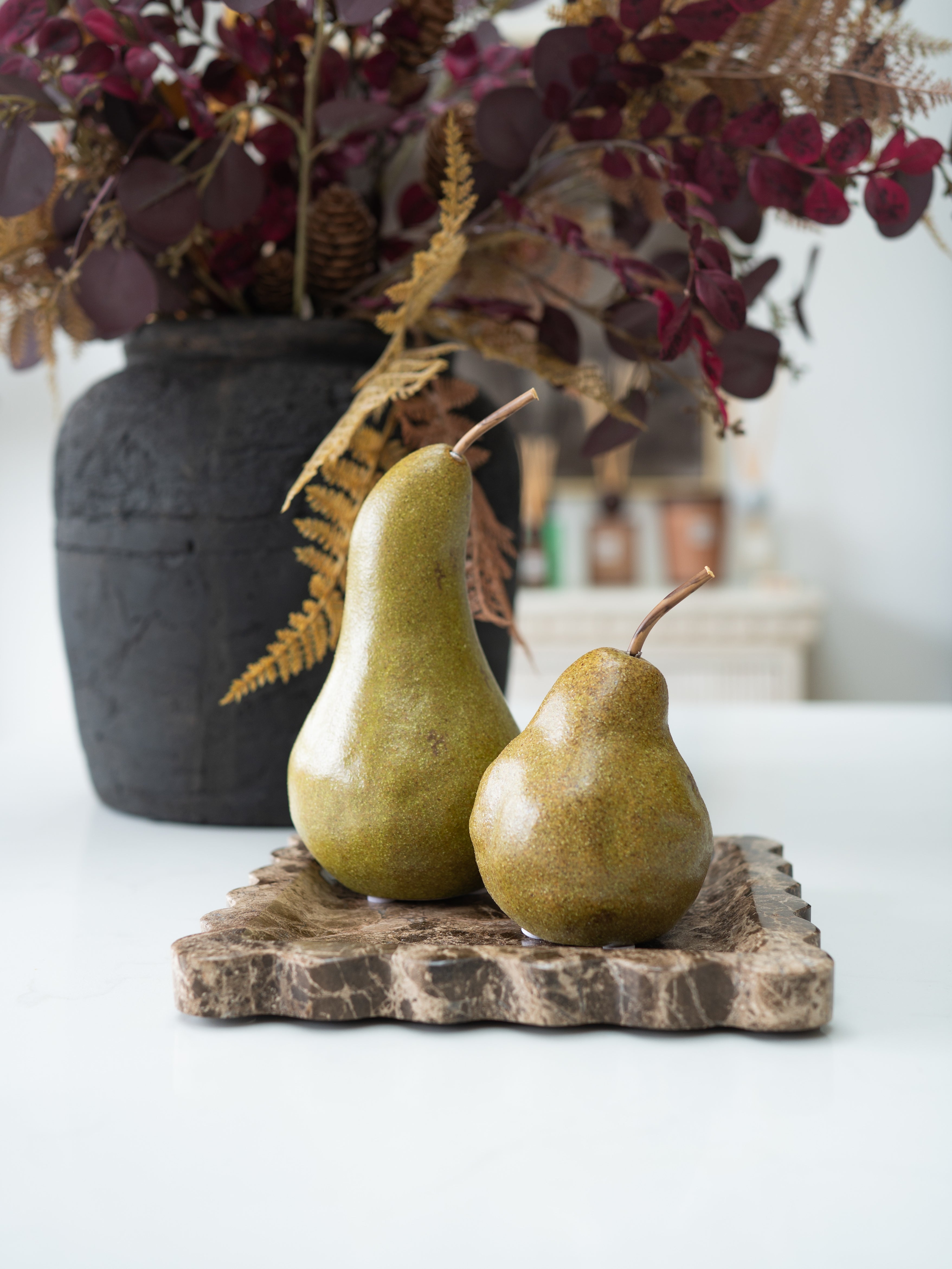 Two decorative pears with a glossy olive-green finish sit on a textured stone tray, with a dark vase of dried foliage in the background.